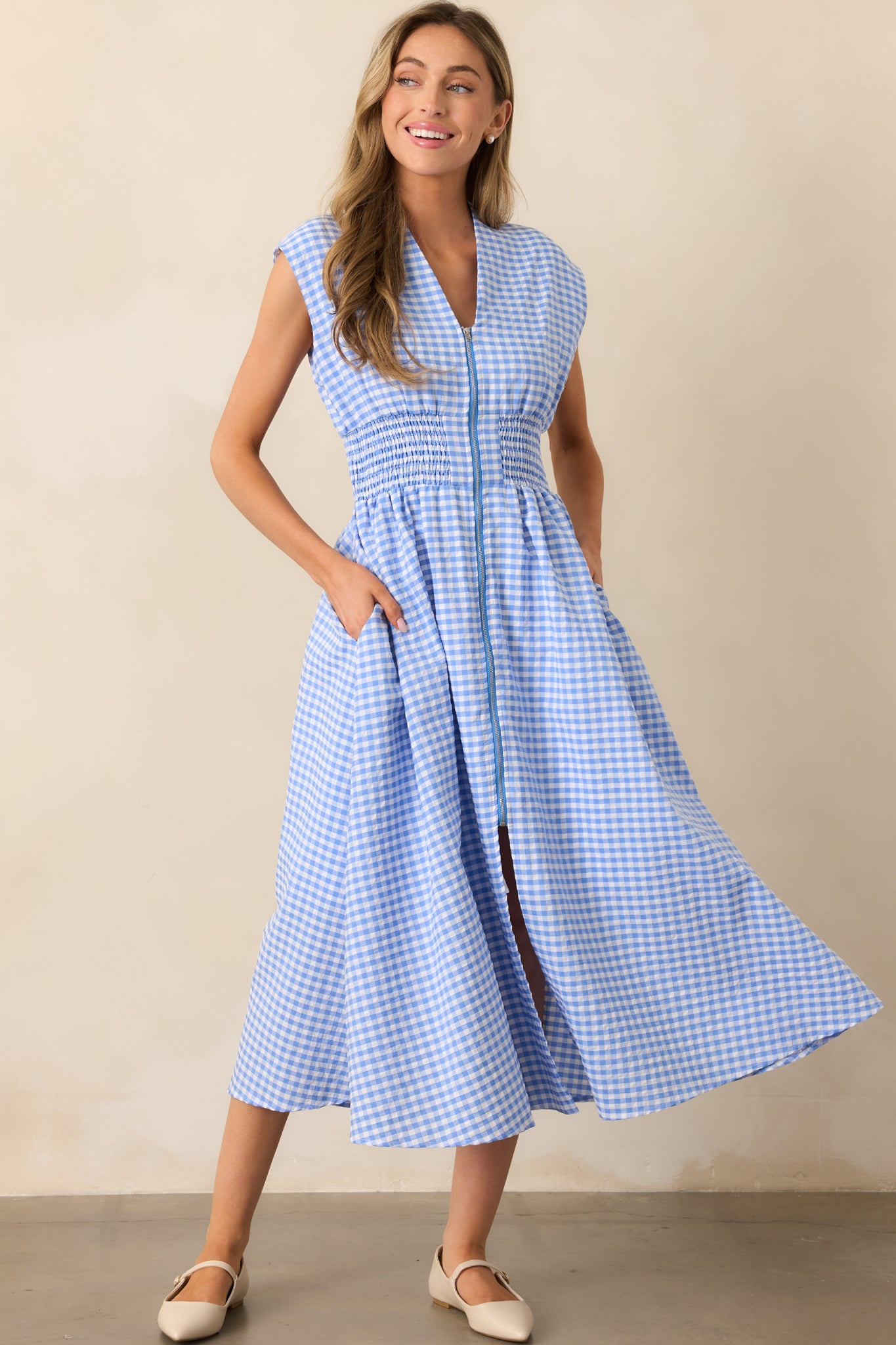 This blue gingham midi dress features a V-neckline, padded shoulders, capped sleeves, a full zipper front, a fully smocked waist, functional hip pockets, and a front slit.