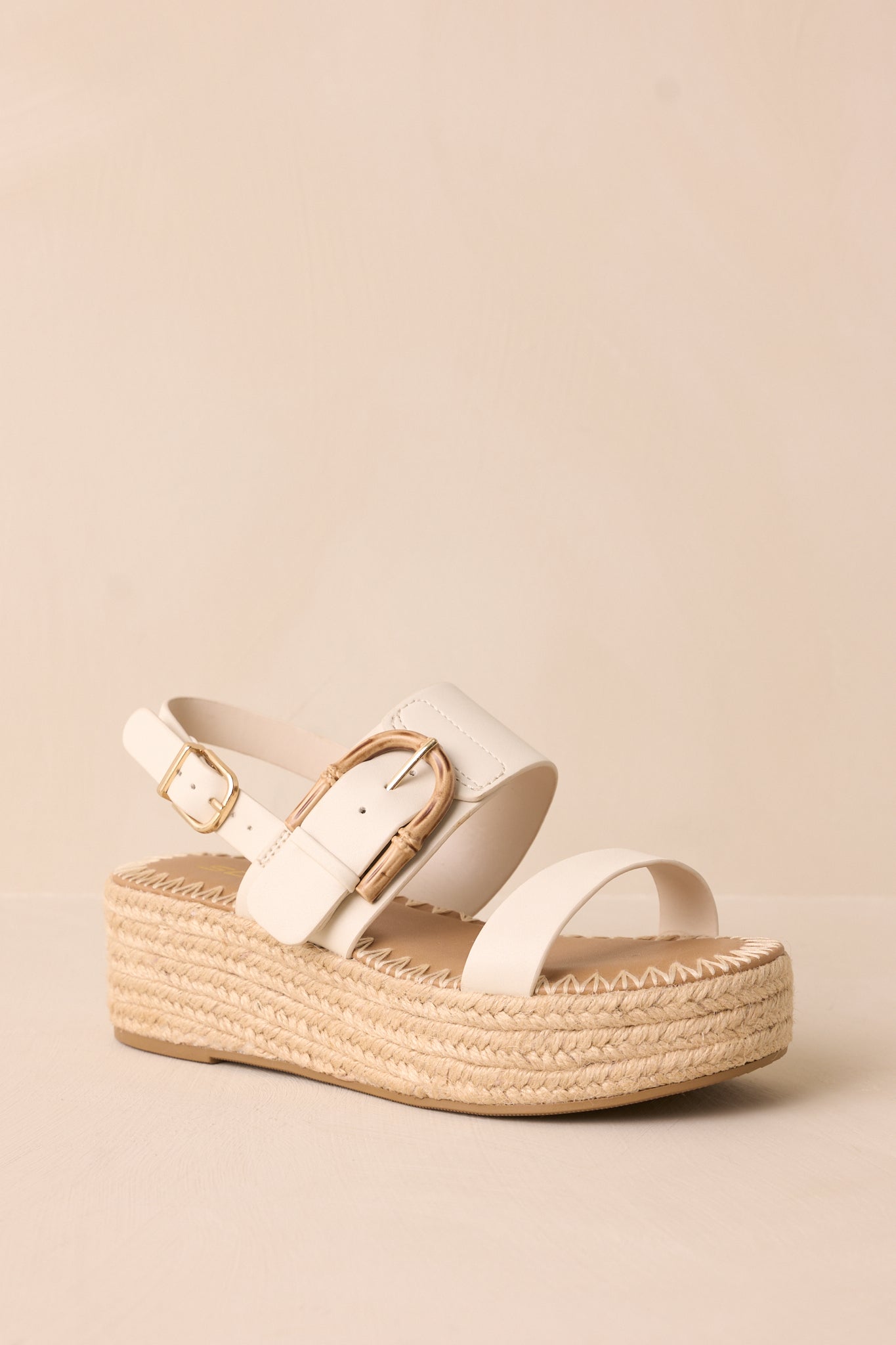  A stylish pair of ivory sandals featuring a rounded toe, ivory toe strap, and a woven straw platform wedge heel, set against a beige background.

