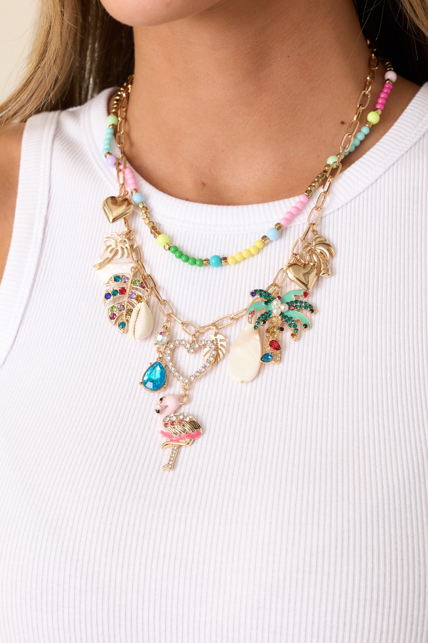 This layered charm necklace features two separate gold chains: a shorter one with pastel beading and a longer one adorned with colorful rhinestone charms, palm trees, hearts, and a seashell charm.