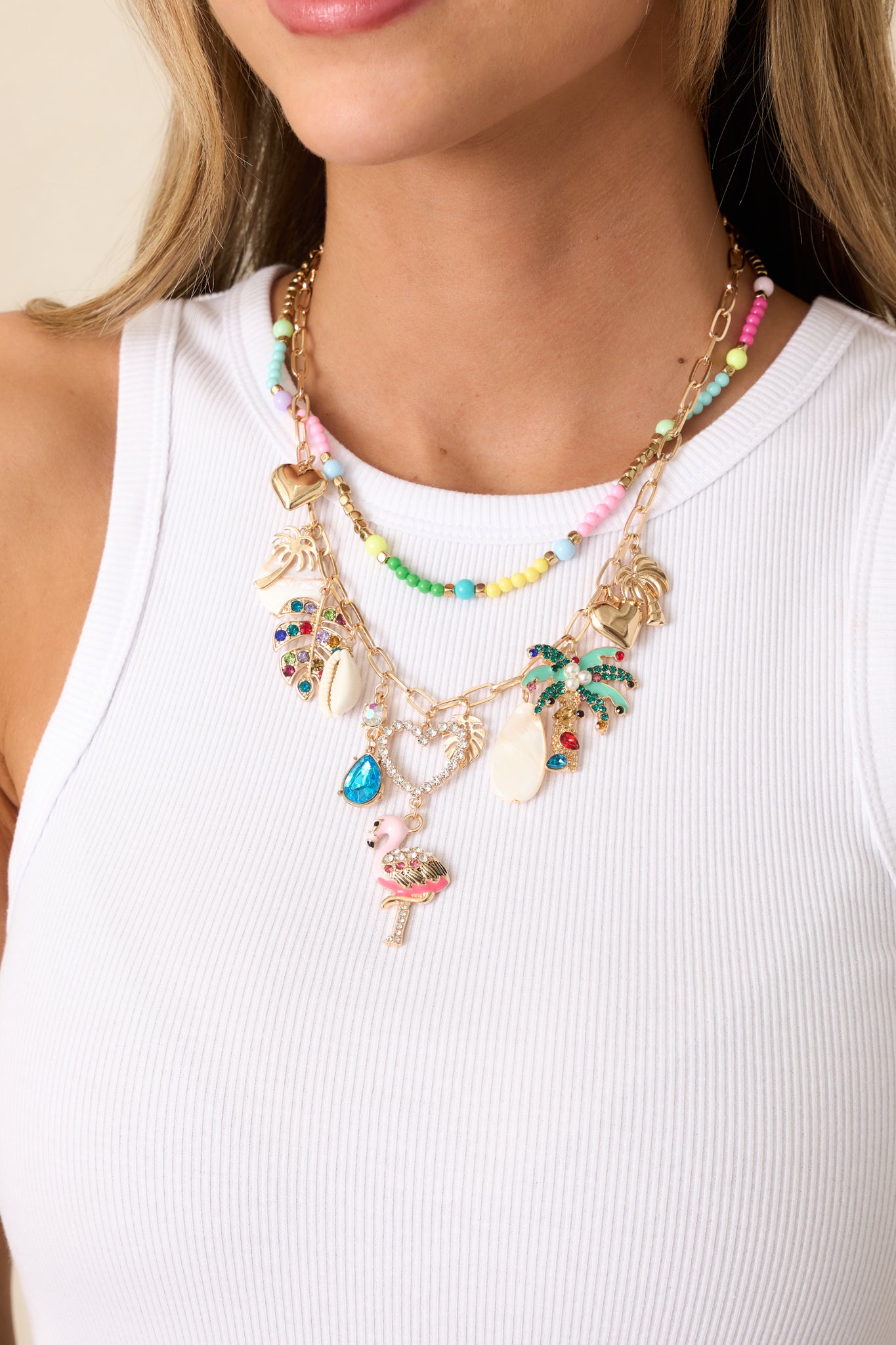 A detailed shot of the gold layered charm necklace, highlighting the intricate beading on the shorter necklace and the variety of beach-themed charms on the longer layer, including palm trees, hearts, and rhinestone accents.