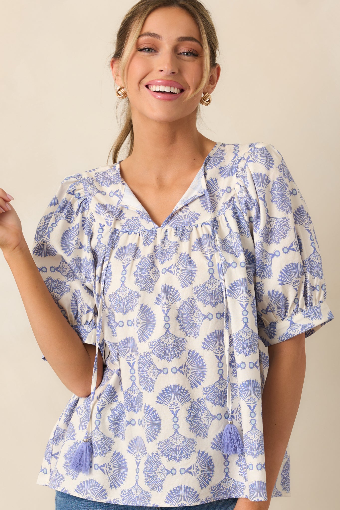 A blue blouse featuring a self-tie neckline with tassel ties, a beautiful print throughout, a relaxed fit, and short puff sleeves.