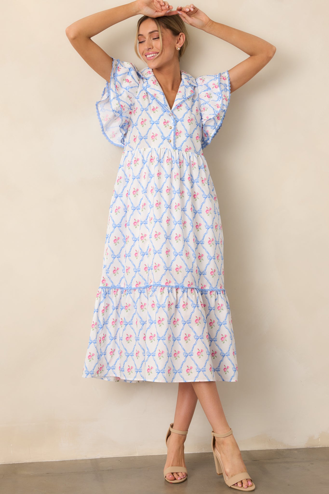 This light blue dress features a collared neckline, button front detailing, and pink trim. The floral and bow print adds charm, with flutter sleeves completing the look.