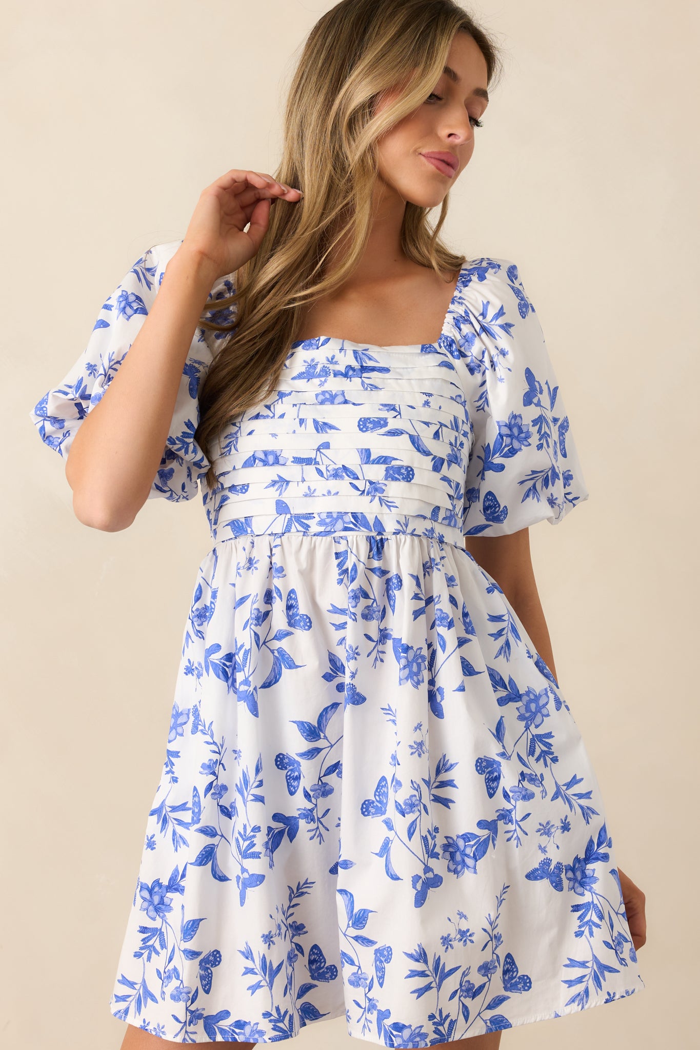 A blue mini dress featuring a straight neckline, fitted pleated bust, relaxed skirt, smocked back, floral print, and puff sleeves.