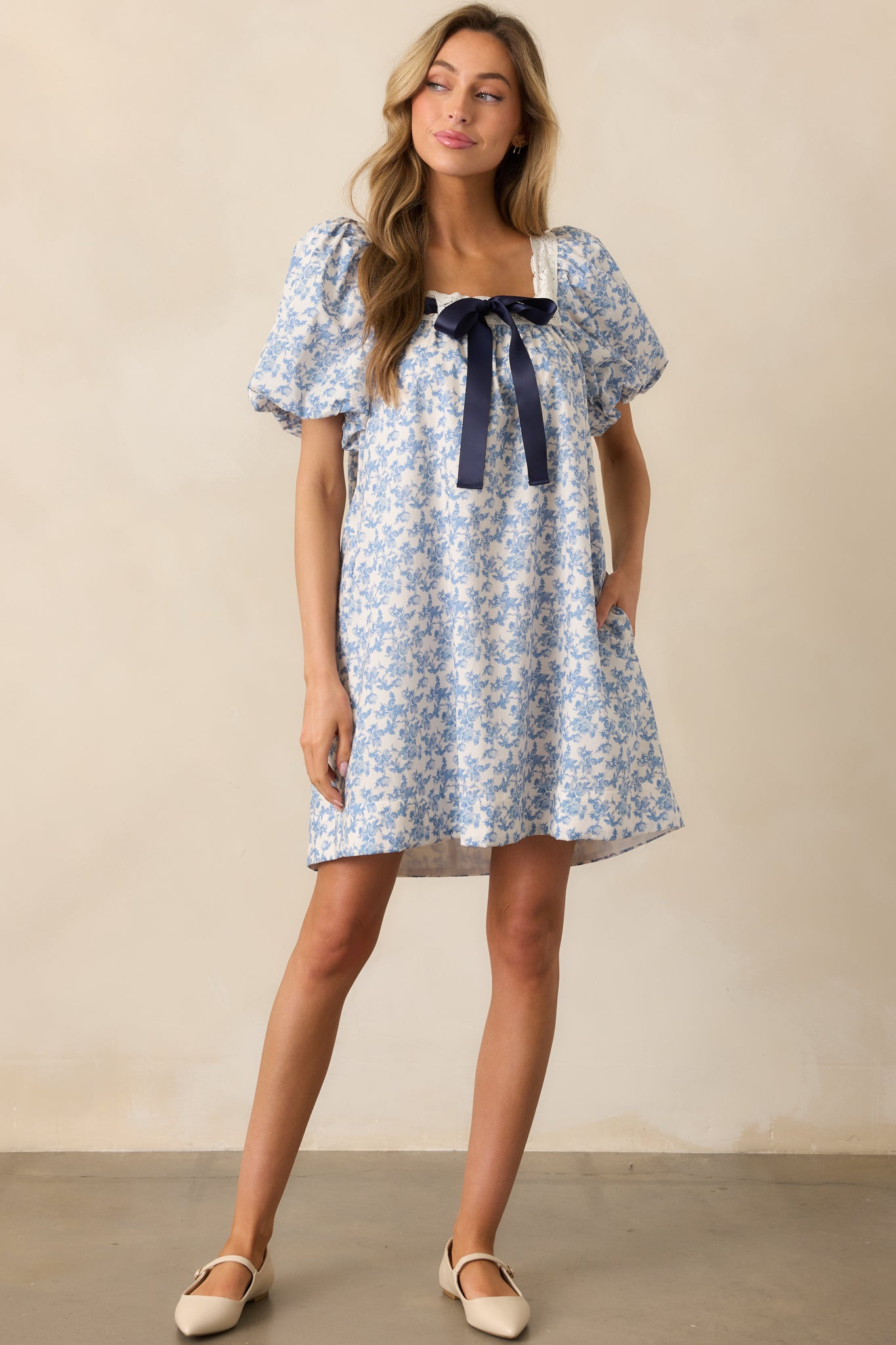  A front-facing shot of the blue mini dress, highlighting the puff sleeves, lace trim detailing, self-tie satin navy bow, square neckline, and the dress's flattering mini length.