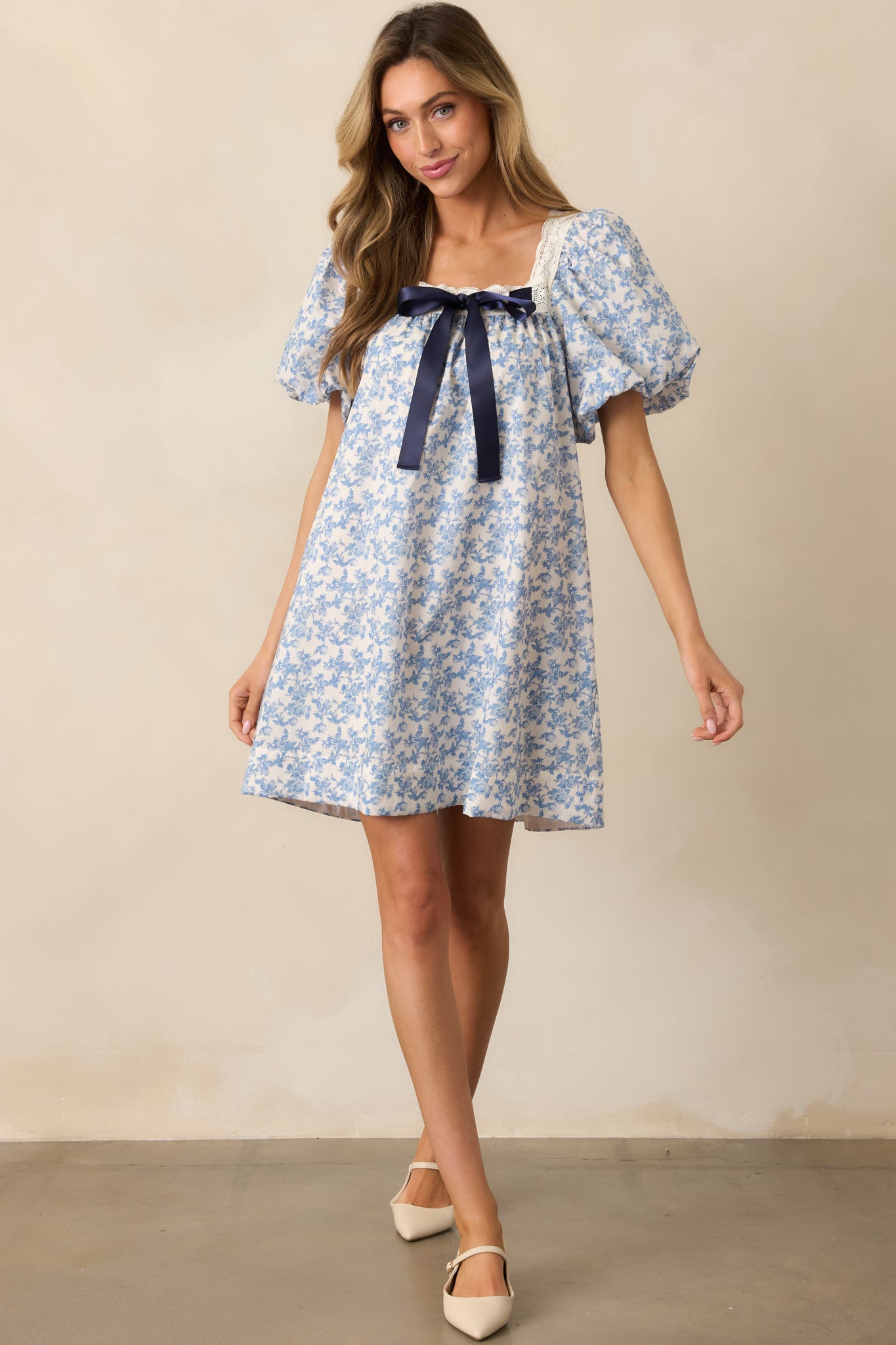 A full-length view of the blue mini dress featuring puff sleeves, lace trim detailing, a self-tie satin navy bow at the waist, and two functional side pockets, creating a playful and feminine look.