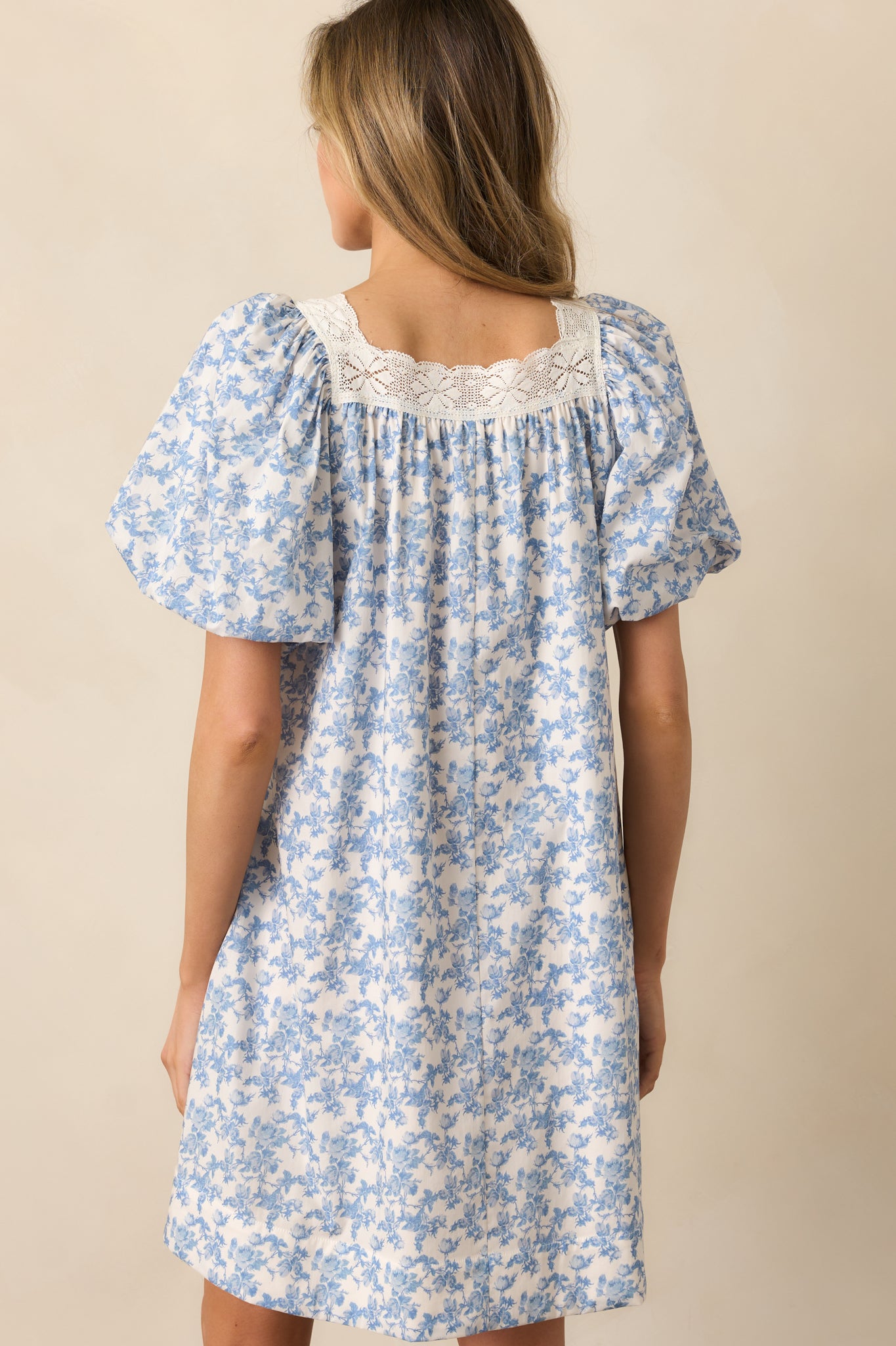 A back view of the blue mini dress, emphasizing the puff sleeves, lace trim detailing, and self-tie satin navy bow at the waist, with the functional side pockets subtly visible.