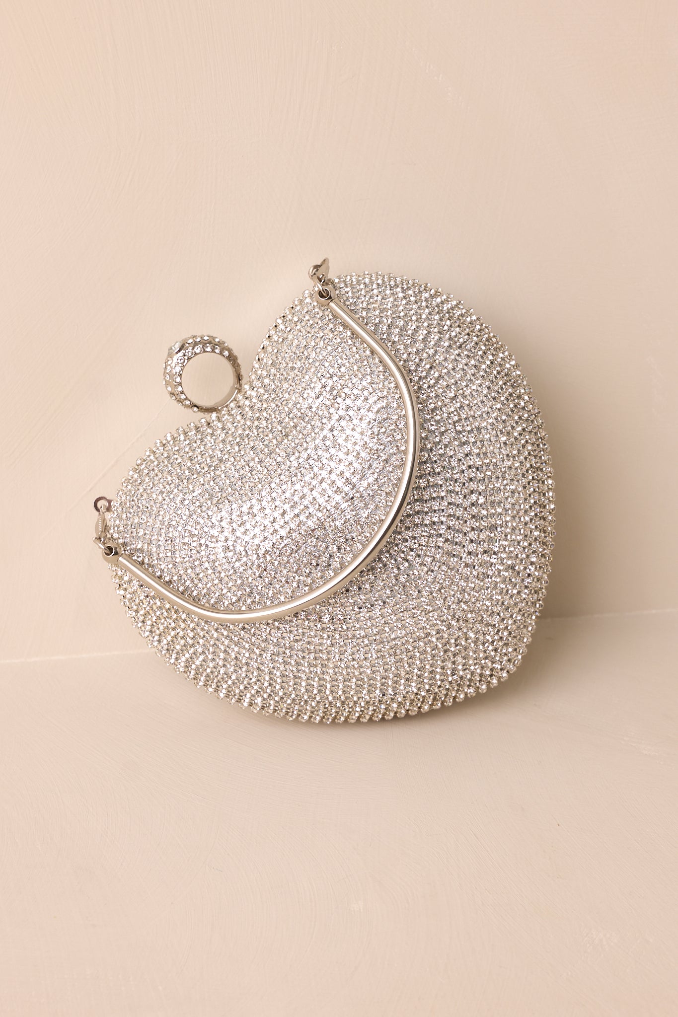 A heart-shaped silver clutch with clear rhinestones on one side, a metallic silver back, and a "ring" style rhinestone closure.