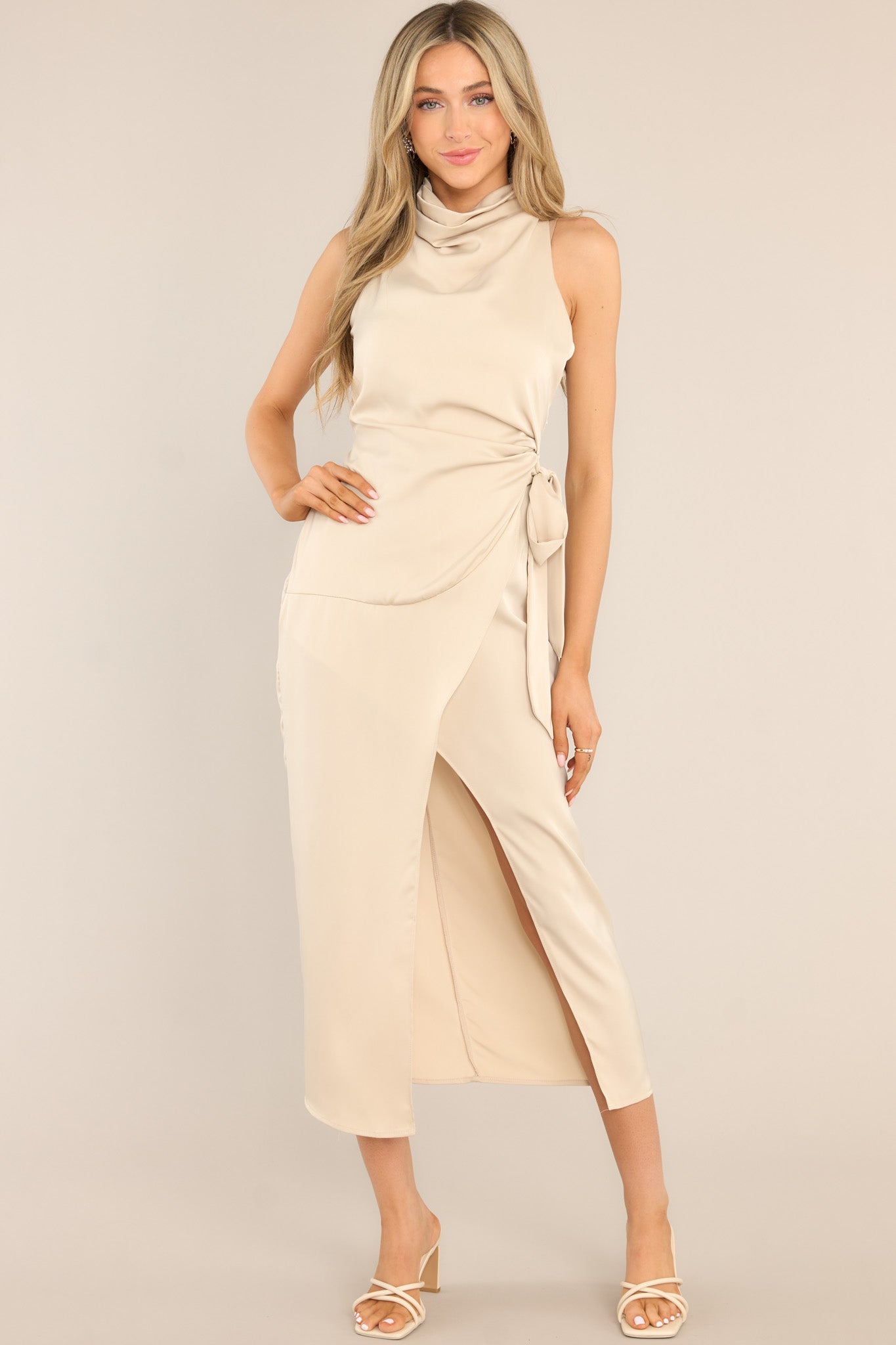 This beige dress features a high cowl neckline, a zipper down the back, a wrap-like design with a self-tie feature at the waist, and a front slit. 