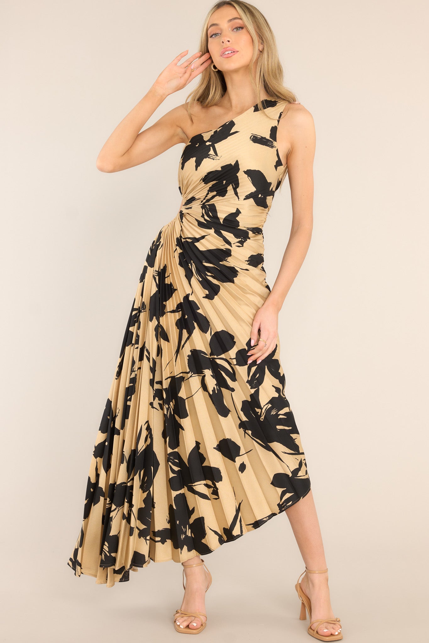 This tan and black dress features an asymmetrical neckline, a side zipper, a circular waist cutout, pleats throughout, and a high-low hemline.