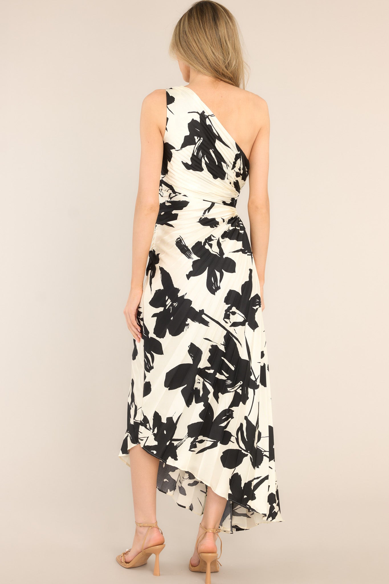 Back view of this dress that features an asymmetrical neckline, a side zipper, a circular waist cutout, pleats throughout, and a high-low hemline.