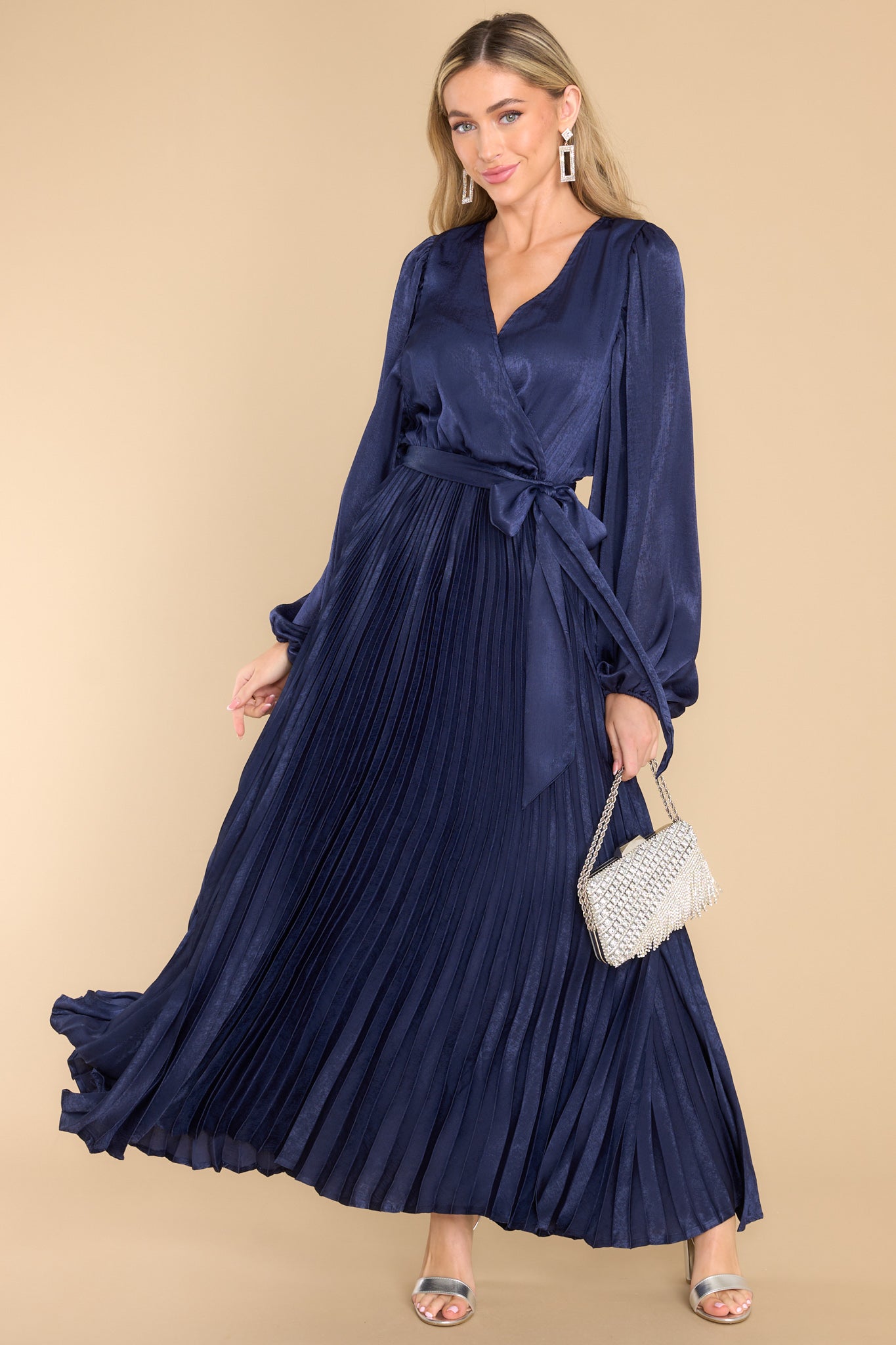 This navy blue dress features a v-neckline with a button clasp, flowy sleeves with elastic cuffs, an elastic waistband, a self-tie, and a pleated skirt. 