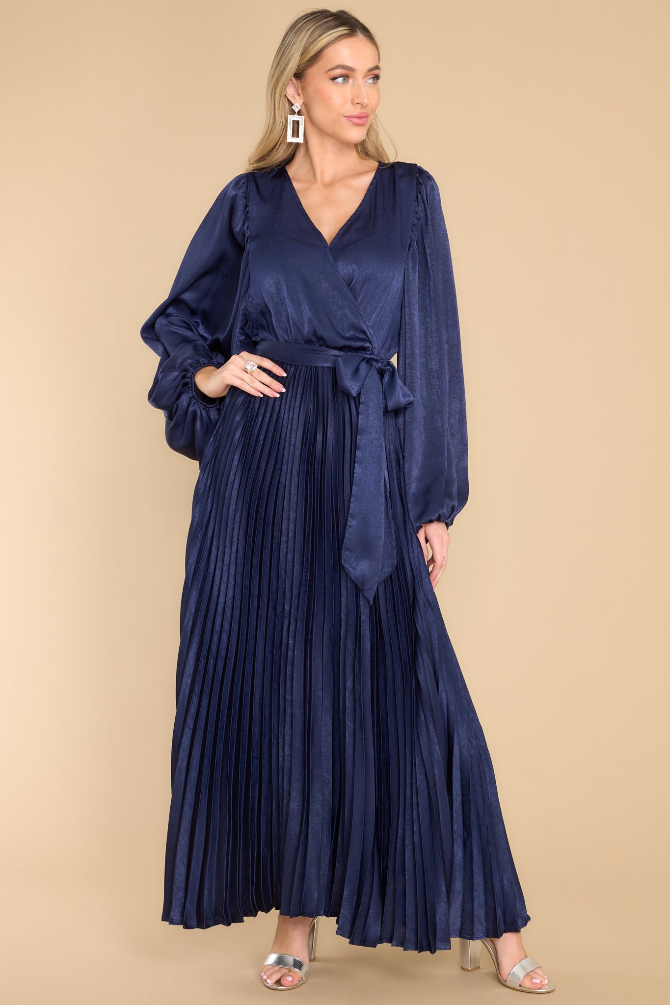 Full body view of  this dress that features a v-neckline with a button clasp, flowy sleeves with elastic cuffs, an elastic waistband, a self-tie, and a pleated skirt. 