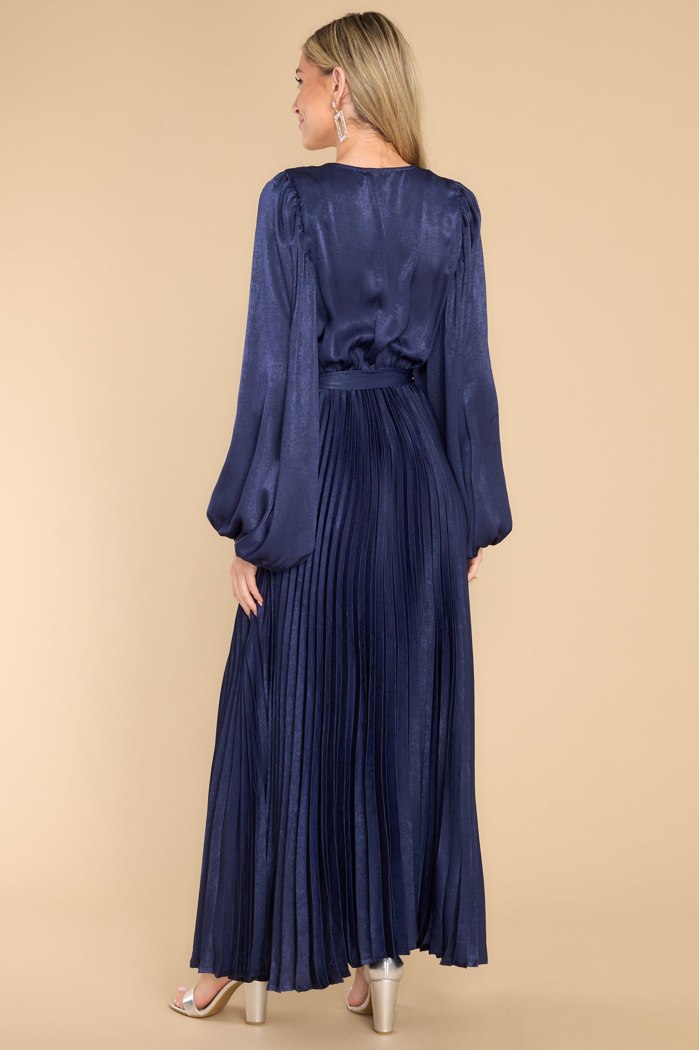 Back view of  this dress that features a v-neckline with a button clasp, flowy sleeves with elastic cuffs, an elastic waistband, a self-tie, and a pleated skirt. 