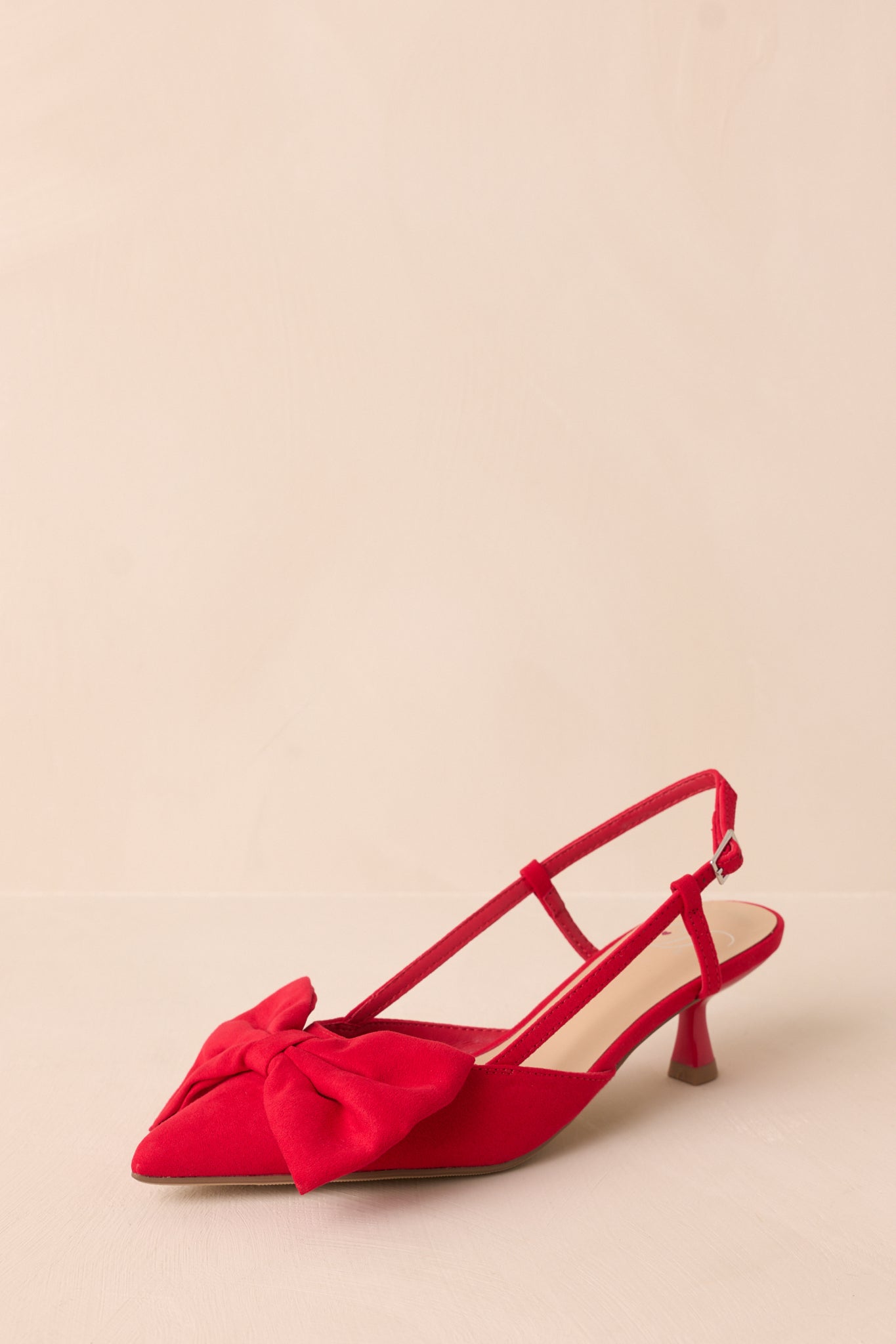 Angled view of red kitten heels with a pointed toe, a large bow on the toe, and an ankle strap featuring a gold buckle, showcasing their elegant shape.