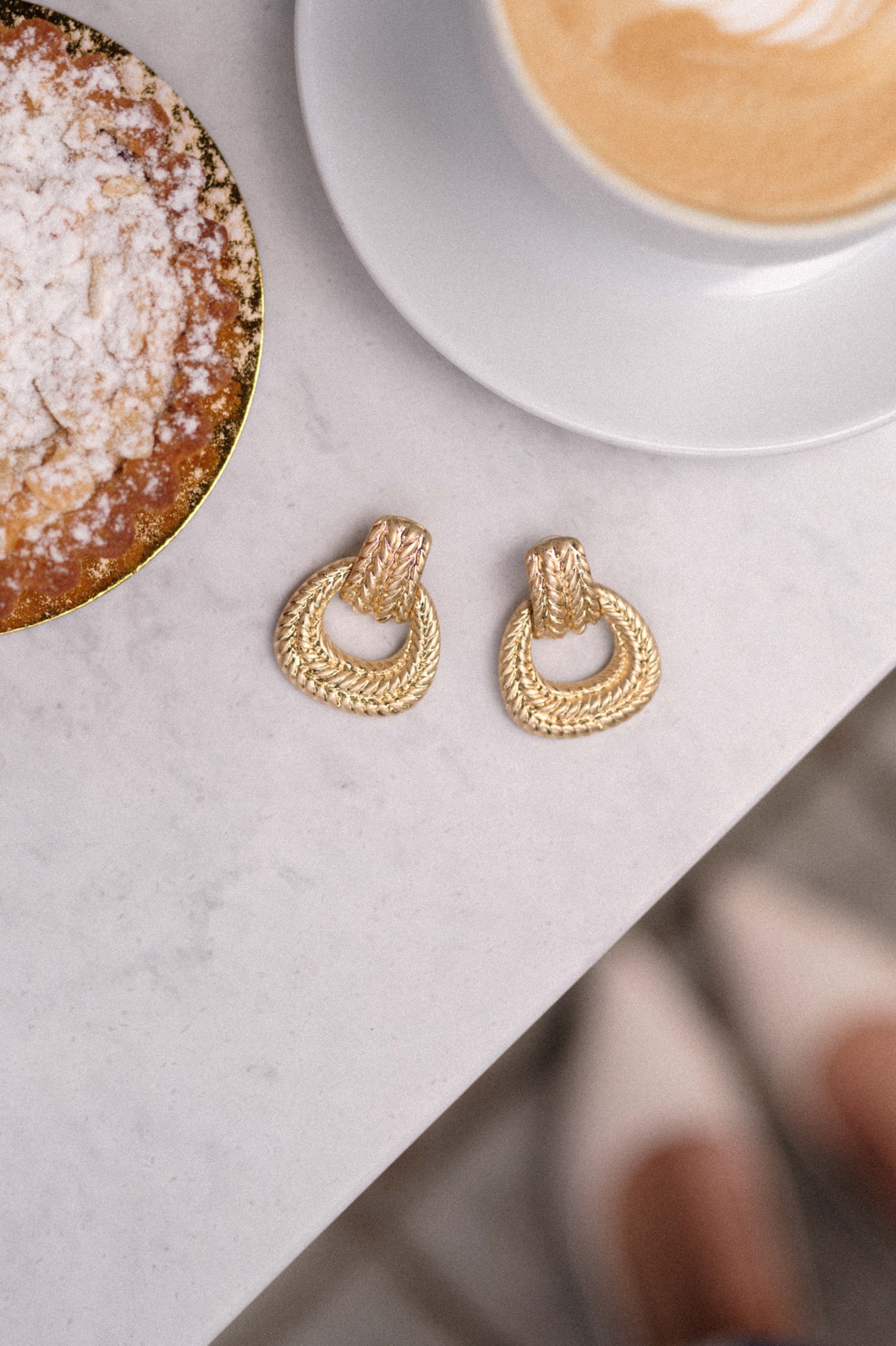 The earrings resting on a table, displaying the braided stud and hoop design against a neutral background, accentuating their elegant appearance.