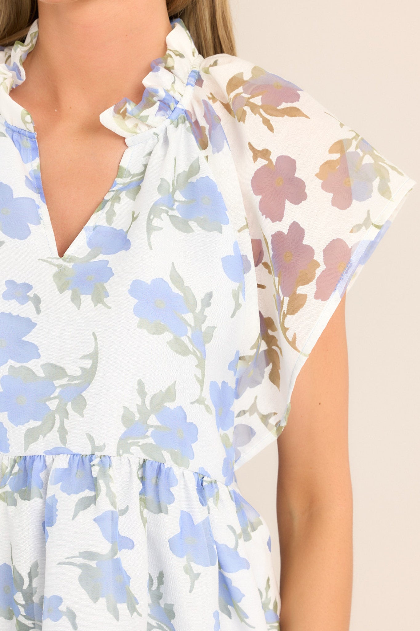 Close up view of this top that features a ruffled v-neckline, a flared waistline, wide transparent sleeves, and a delicate floral pattern.