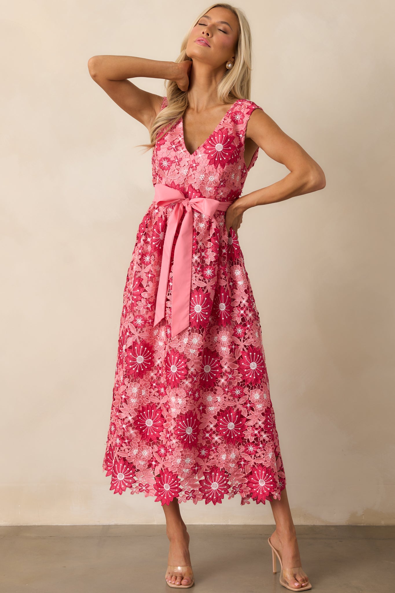 A pink dress with a v-neckline, floral embroidery detail, and a self-tie ribbon waist, featuring a sleek, sleeveless design and midi length.