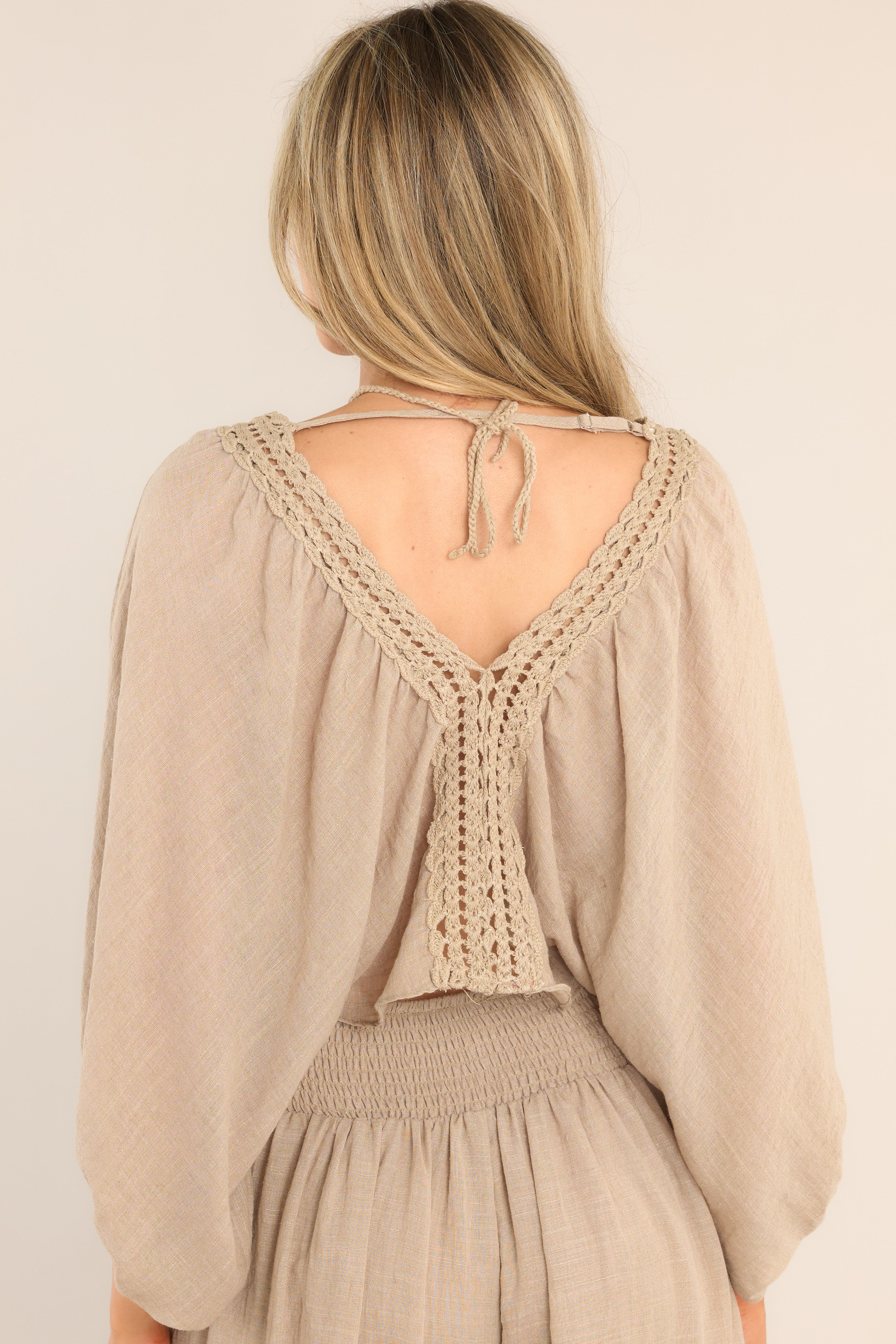 Back view of this top that features a v-neckline, crochet detailing down the middle of the bodice, a self tie feature at the back of the neck, an adjustable strap, a linen like material, and elastic cuffed sleeves.