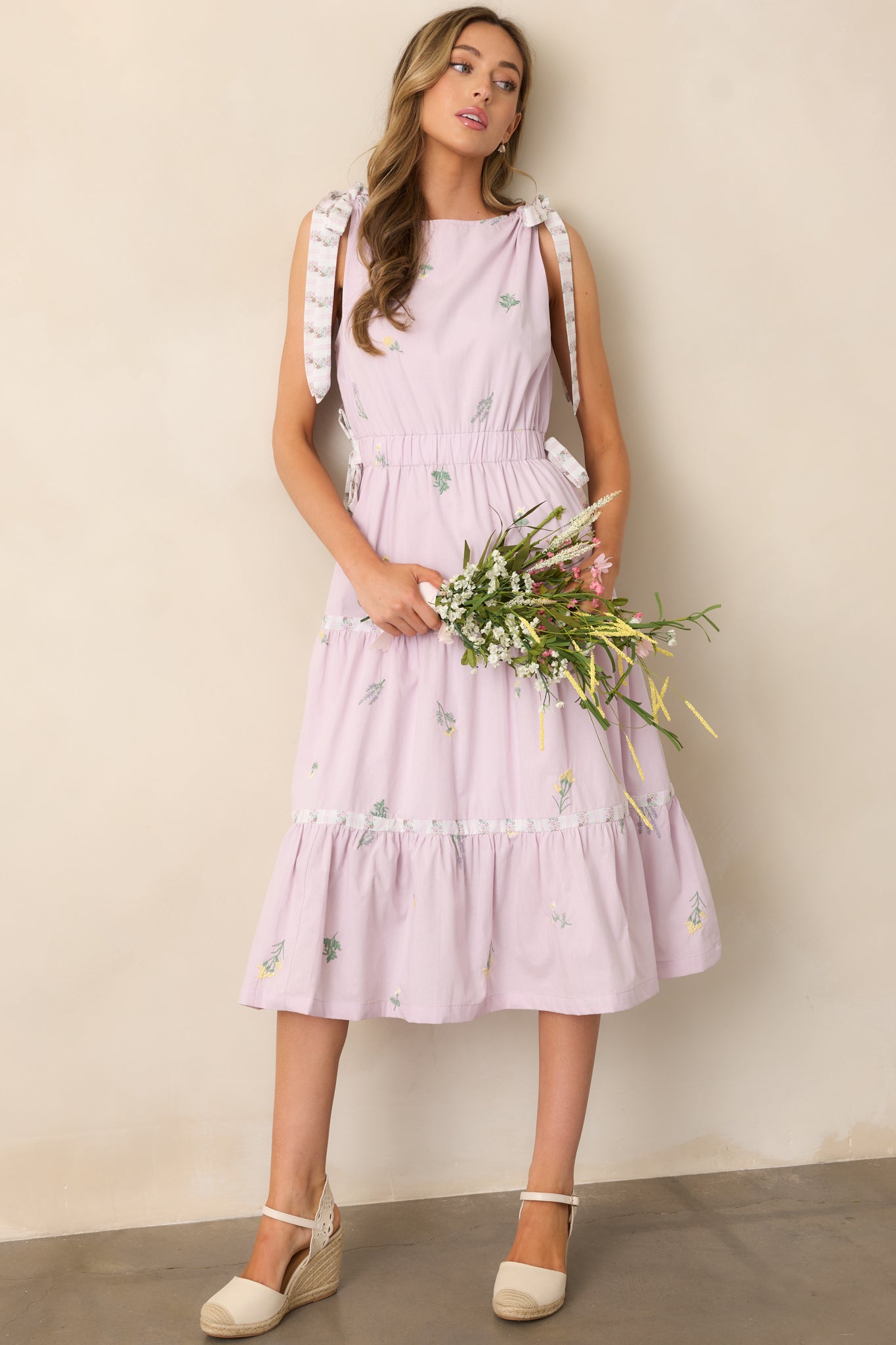 A lavender dress featuring a high rounded neckline, bow shoulder details, an elastic waistband with bow detailing, and an embroidered floral print.