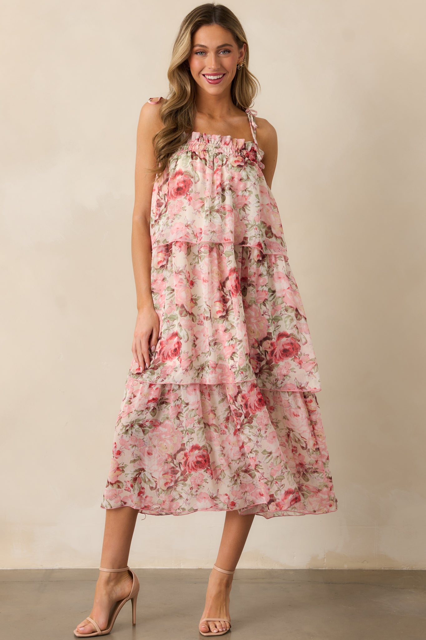 A pink floral midi dress with a tiered design, smocked neckline, and adjustable self-tie shoulder straps, shown from the front. 