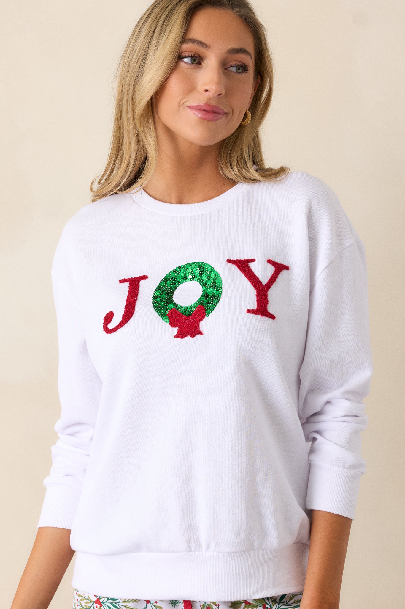 Front view of a white sweatshirt featuring a crew neckline, Joy detailing with a wreath accent, a relaxed fit, and long sleeves with ribbed hems.