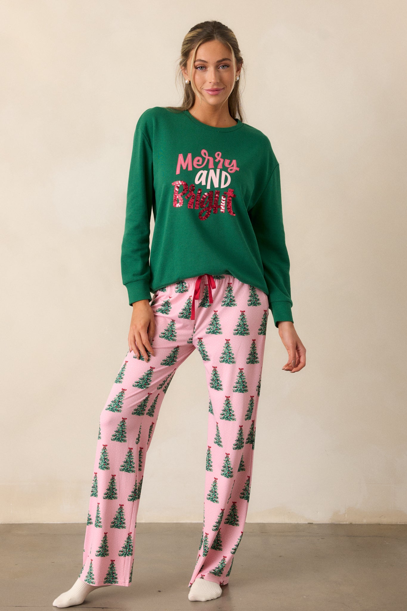 Front view of the pink pajama pants, showcasing the drawstring waistband with red string detailing and the full-length pant legs adorned with a festive green Christmas tree pattern.