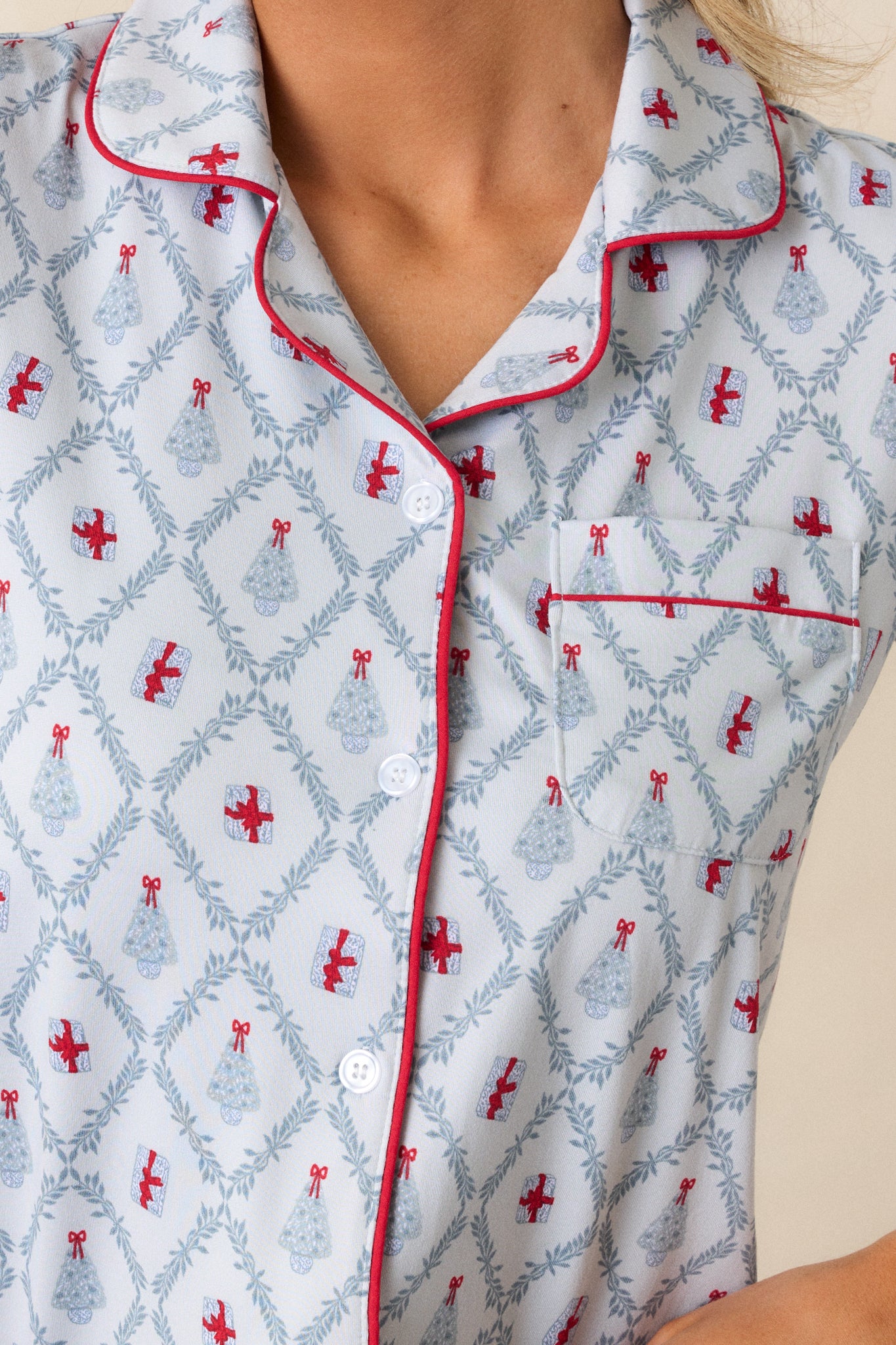 Close up of the breast pocket, functional buttons, and red trimmed collar on this button up, light blue pajama top featuring a tree and present holiday print. 