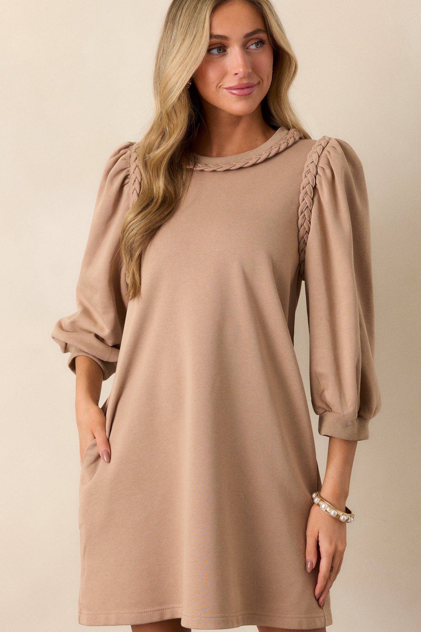 Tan mini dress featuring a braided crew neckline, braided detailing at the shoulders, ribbed cuffs, a box fit, and 3/4 sleeves.