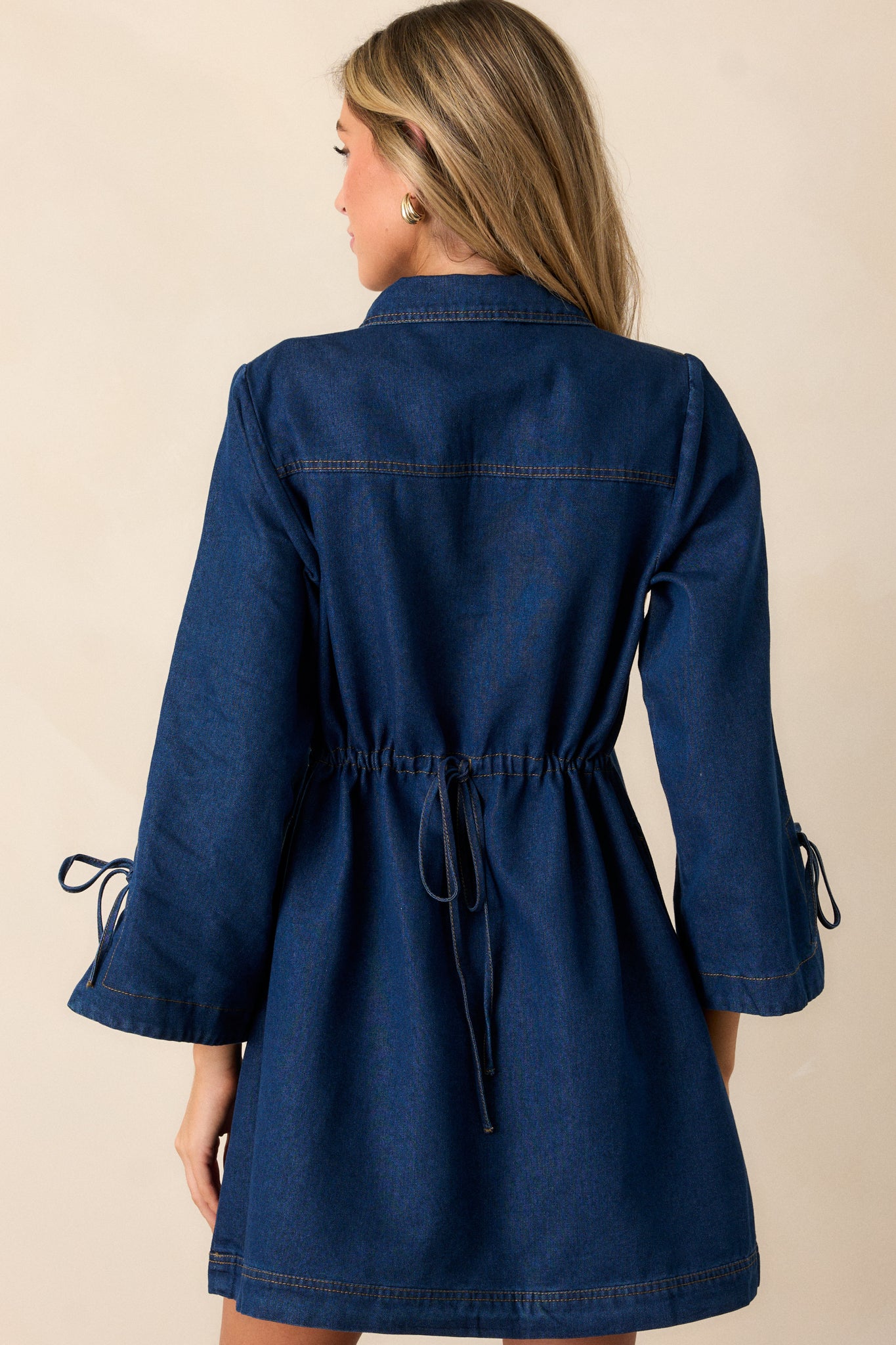 A back view of the dark wash denim dress, emphasizing the tie cinch detail and the flow of the long sleeves with slit openings.