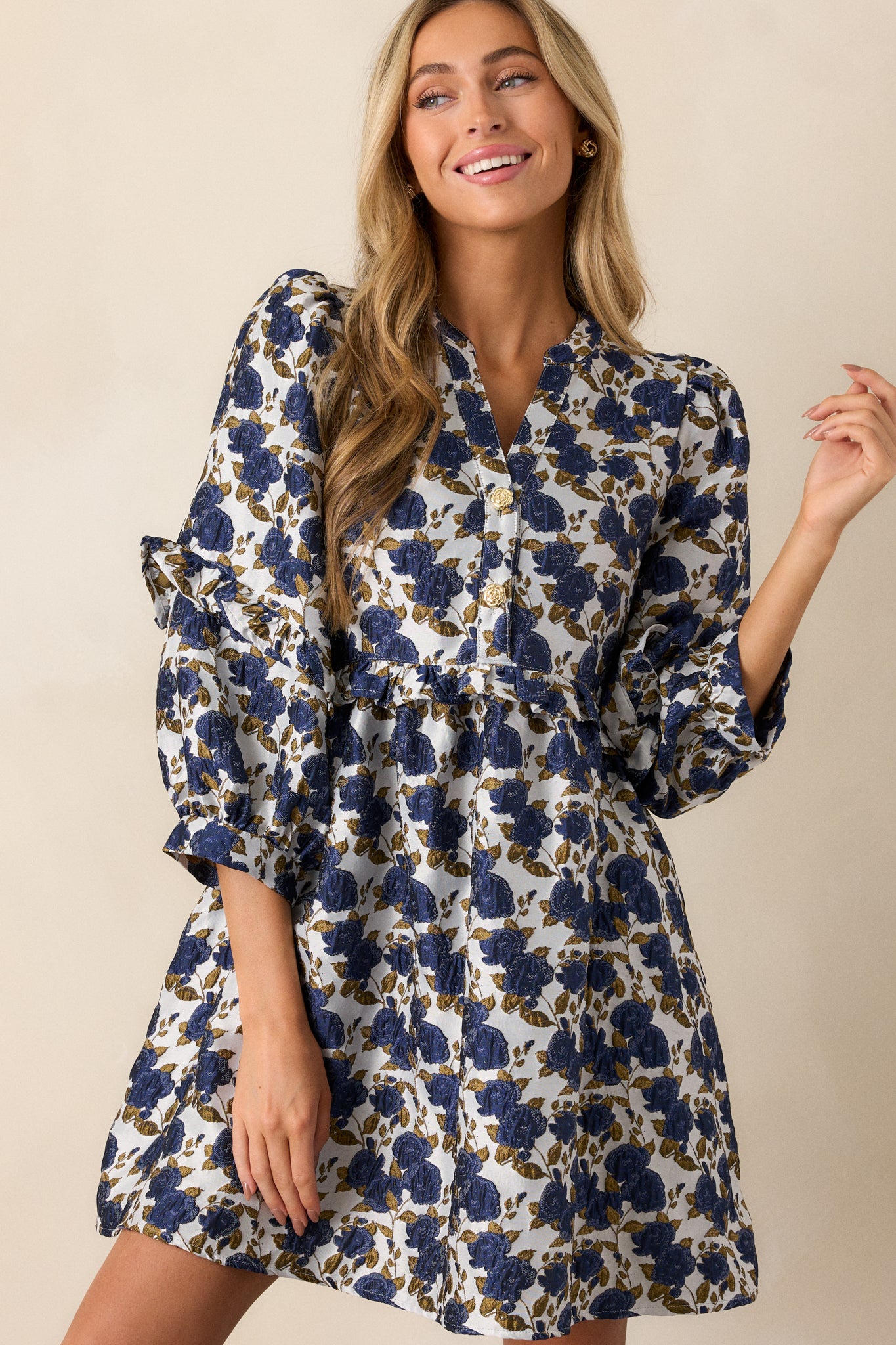 Full view of a navy dress featuring floral pattern, a slight v-neckline with functional gold floral embellished buttons. a ruffle waistline, and 3/4 length puff sleeves with ruffle detailing.