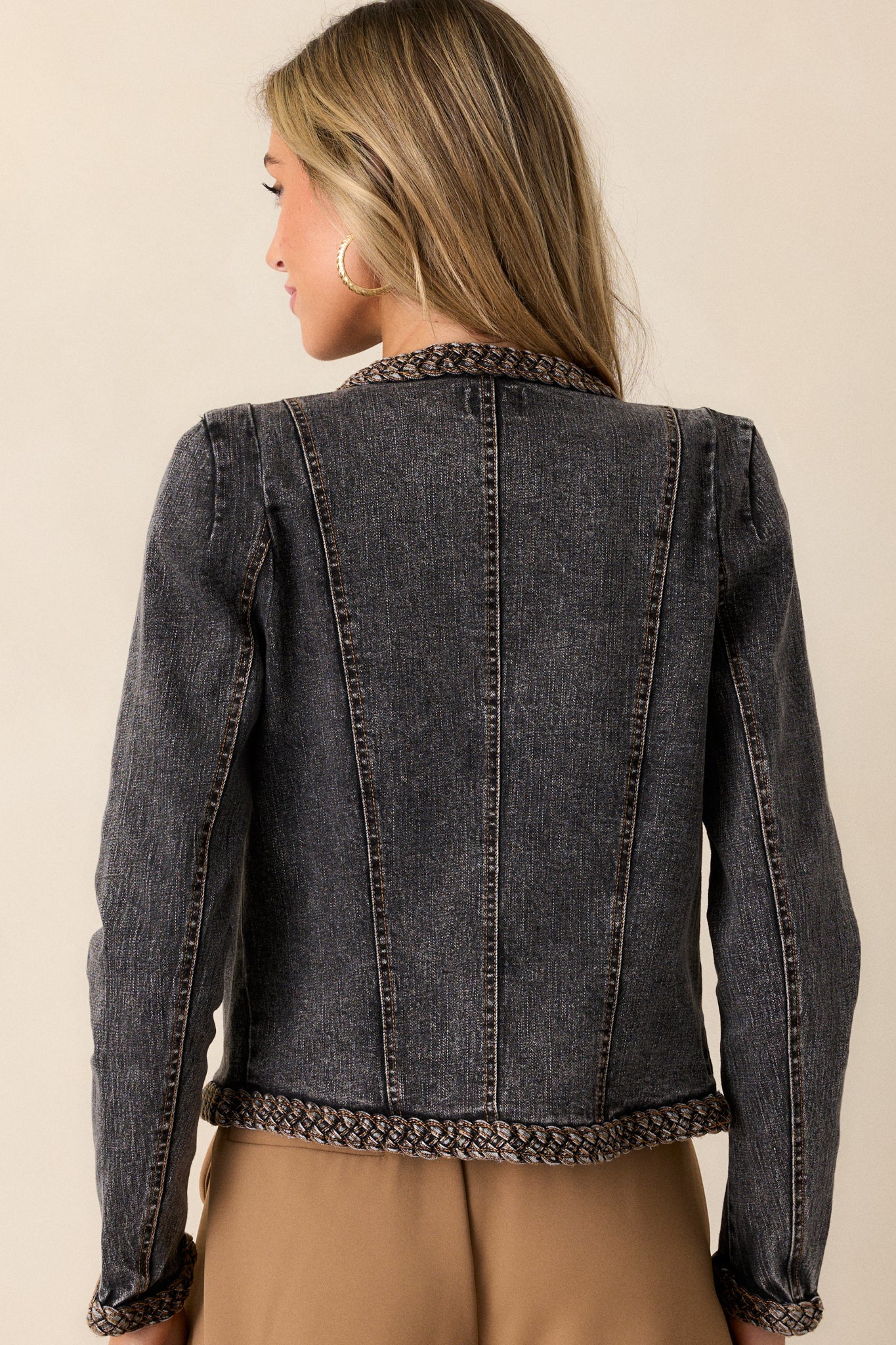 A back view of the black denim jacket, showcasing the long sleeves, braided seam accents, and the contrast of the brown stitching throughout.