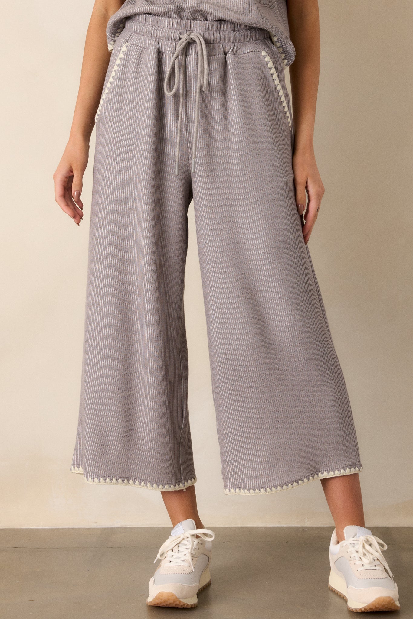 Close-up of these grey pants that feature an elastic waistband, an optional waist drawstring, an ivory ruffle trim, functional hip pockets, a soft stitched material and a wide leg