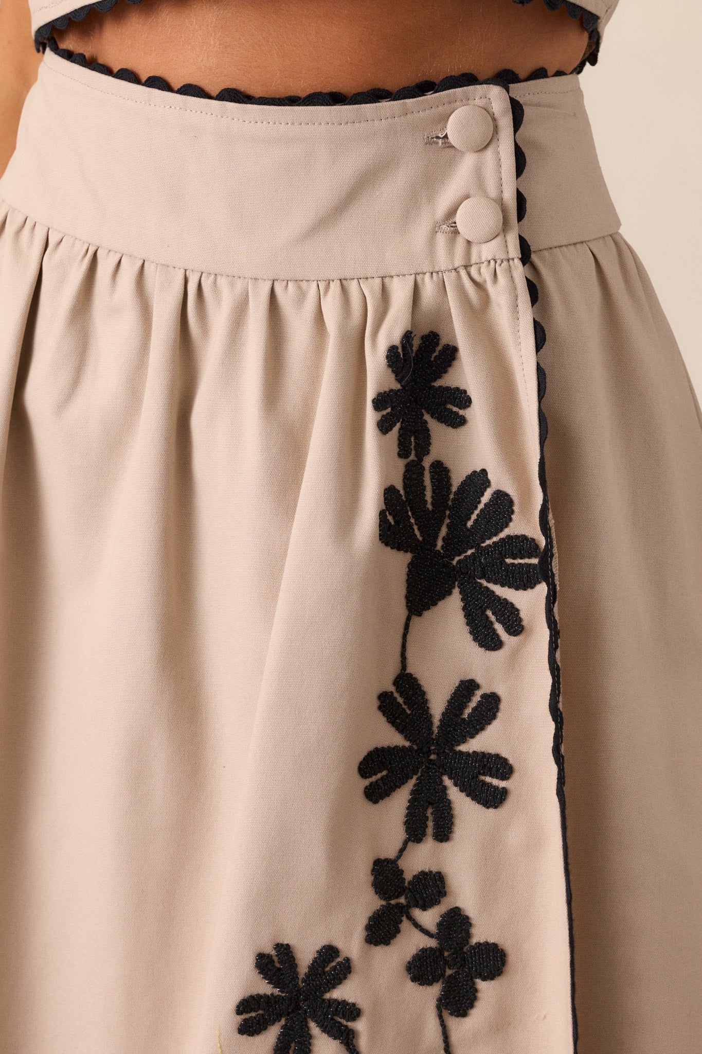 An extreme close-up of the taupe skirt, showcasing the texture of the fabric, the delicate black floral embroidery, and the neatly stitched ric rac trim.