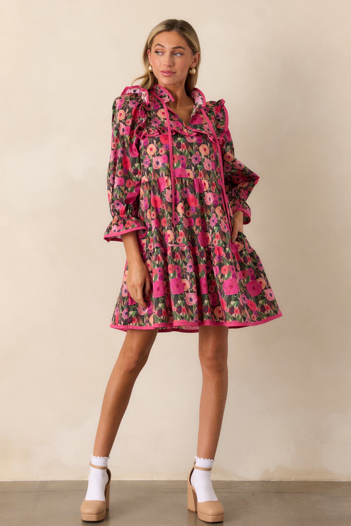 A full-length view of a pink mini dress with a ruffled collar and tie accent, featuring a floral print and functional hip pockets.