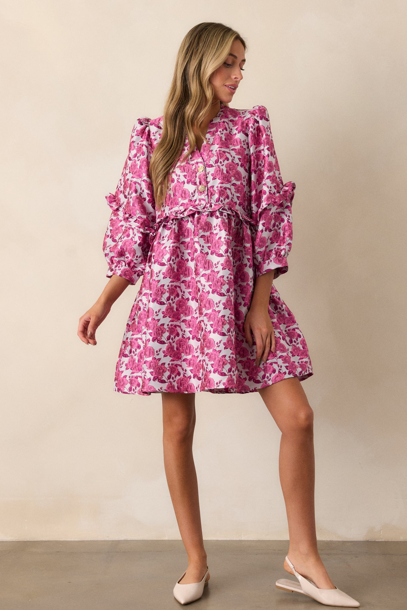 This pink dress features floral pattern, a slight v-neckline with functional gold floral embellished buttons. a ruffle waistline, and 3/4 length puff sleeves with ruffle detailing.