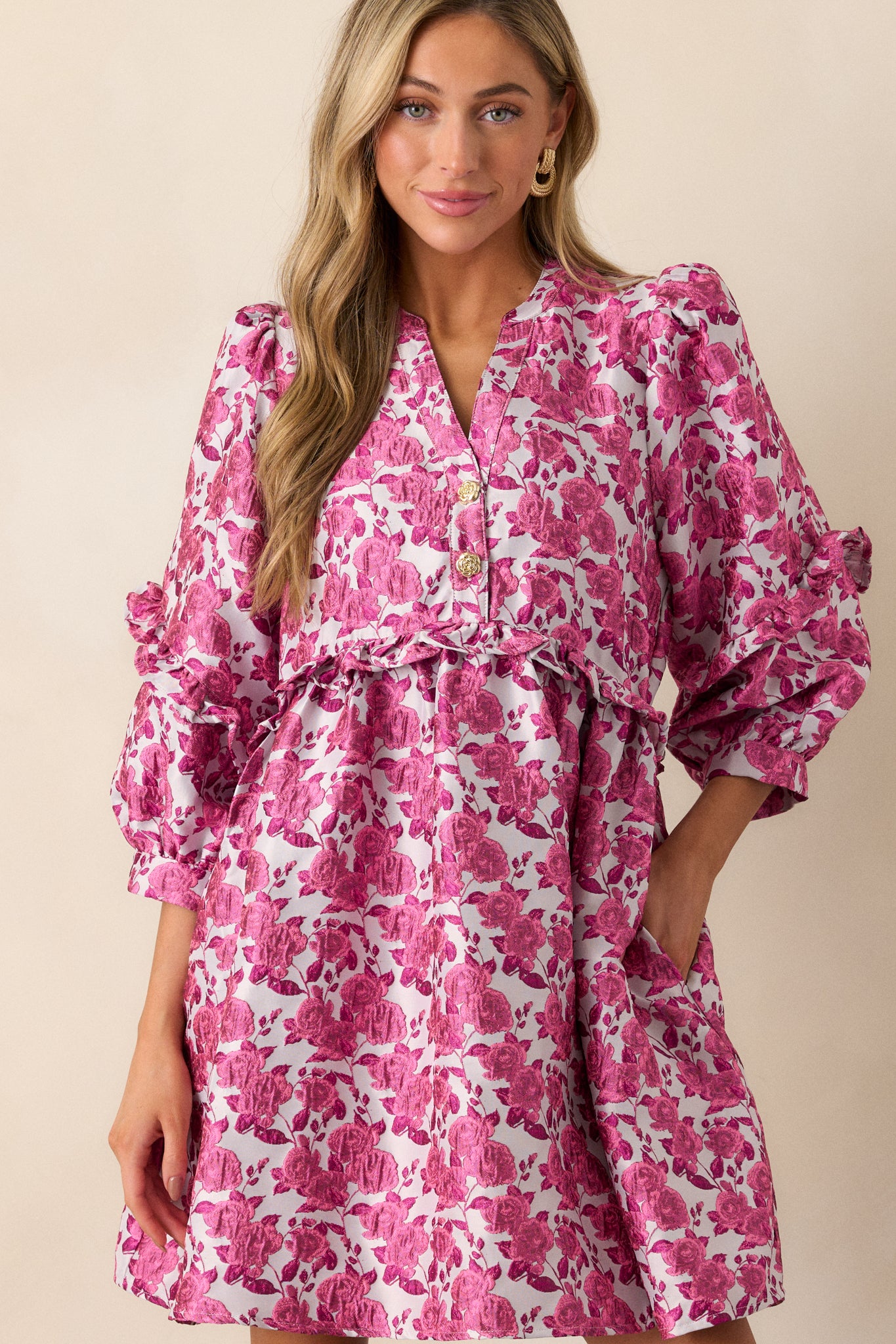 Full view of a pink dress featuring floral pattern, a slight v-neckline with functional gold floral embellished buttons. a ruffle waistline, and 3/4 length puff sleeves with ruffle detailing.