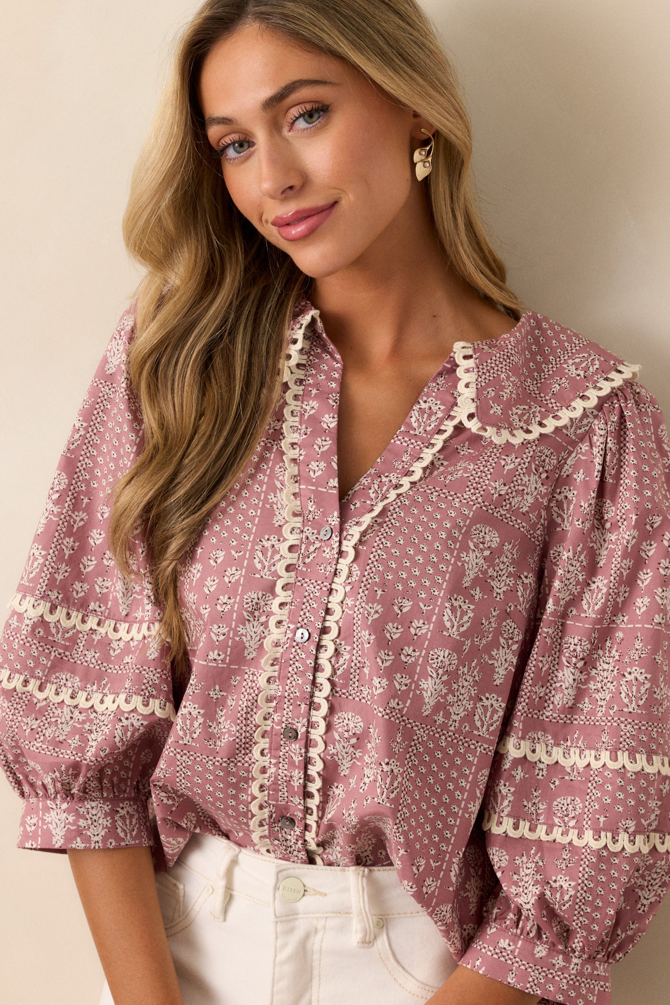 Front, tucked in view of a dusty pink blouse featuring a collared v-neckline, button up front, small scalloped hemming, a beautiful floral pattern, and 3/4 puff sleeves with scalloped detailing.