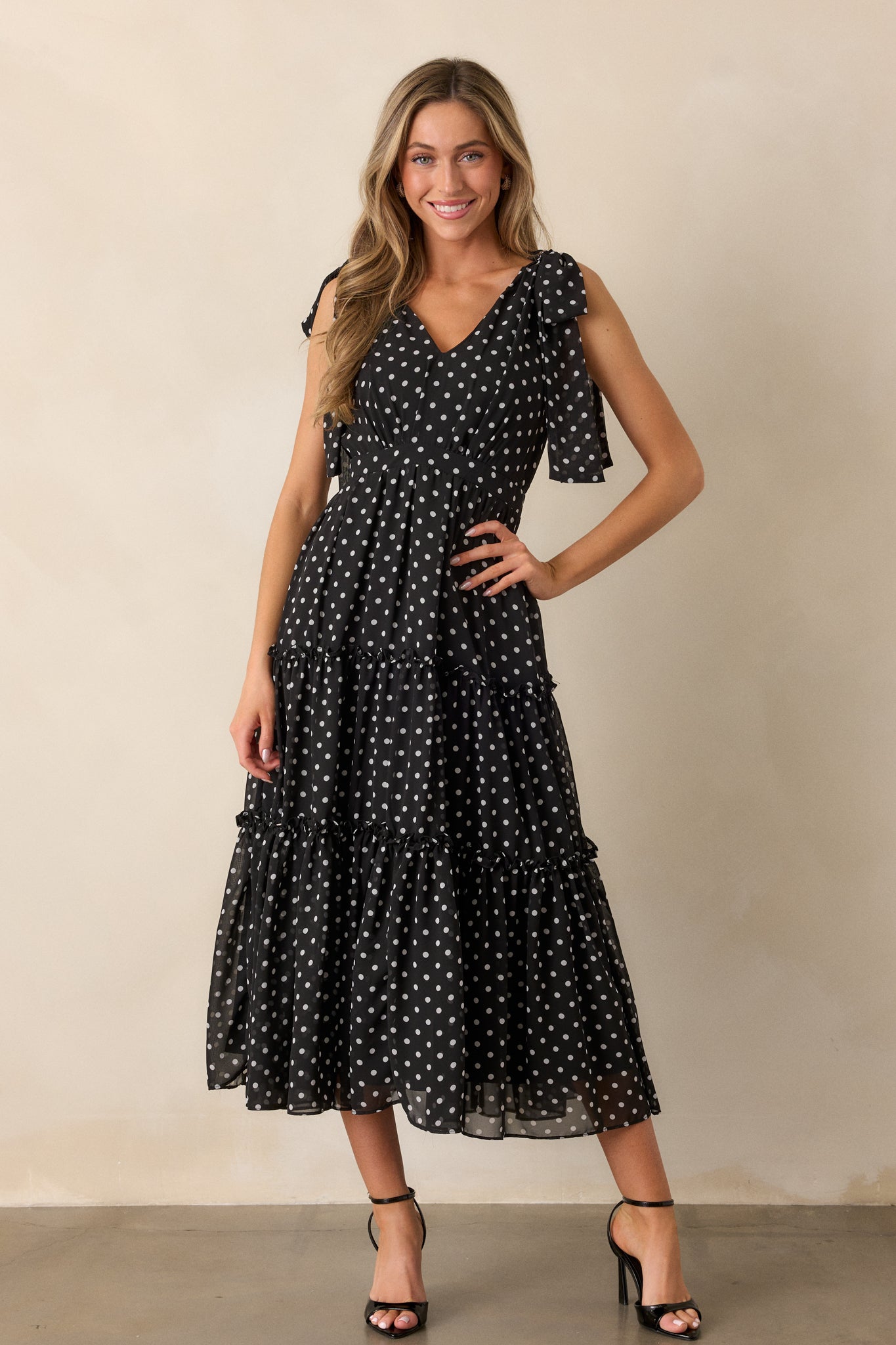 Full view of the black dress featuring a v-neckline, tiered skirt with ruffled hems, and self-tie sleeves. The dress has a playful polka dot print and a discrete back zipper.