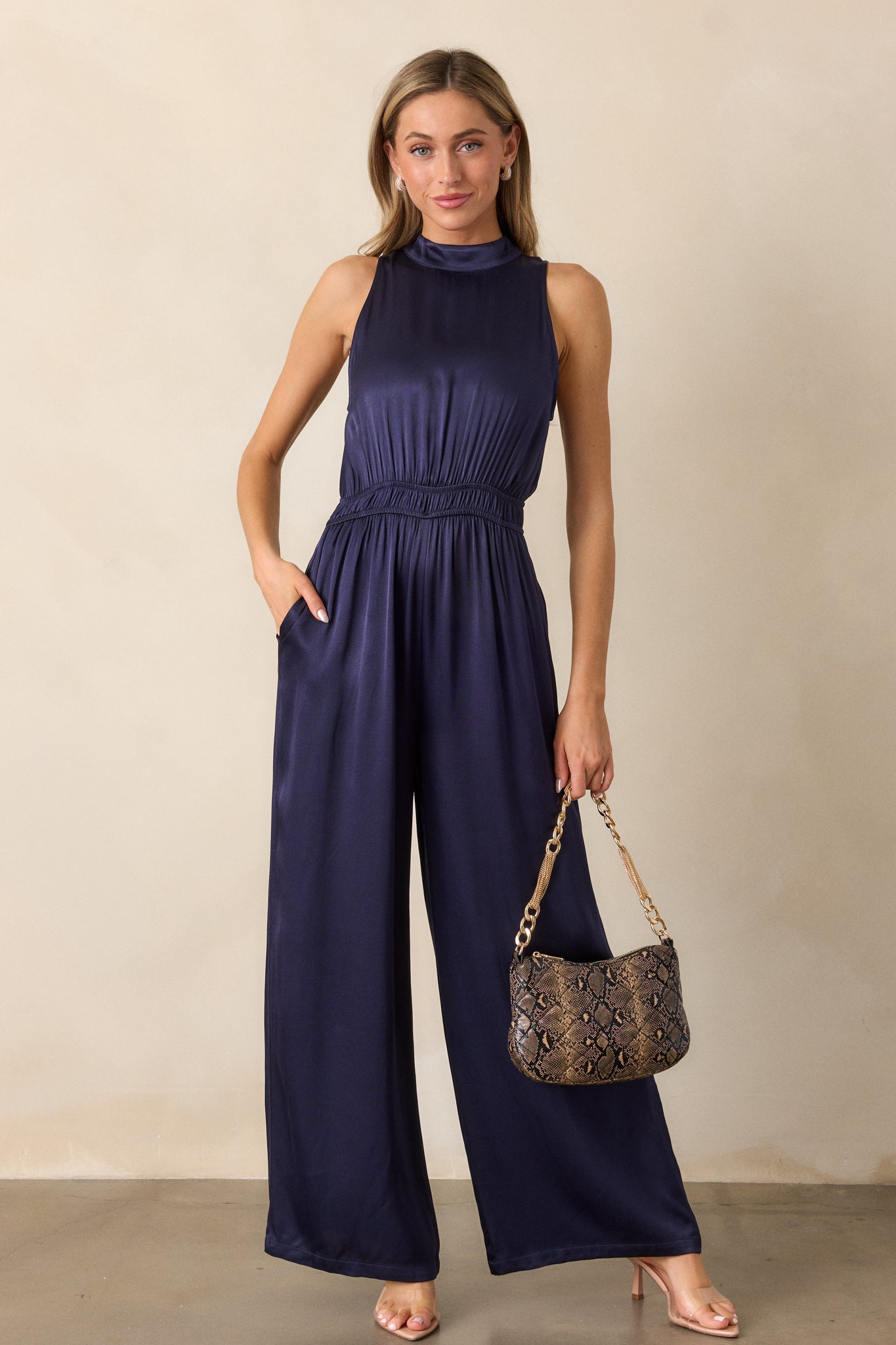 Full front view of the midnight blue jumpsuit, featuring a high neckline with a button neck clasp, cinched waist with elastic detailing, and long flowy pant legs.