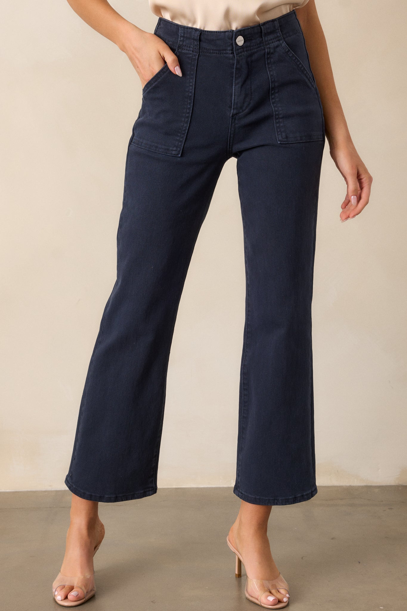 A closer shot of the waist area of the navy jeans, focusing on the button and zipper closure, along with the large front pockets. The high-waisted fit and smooth fabric texture are also visible.