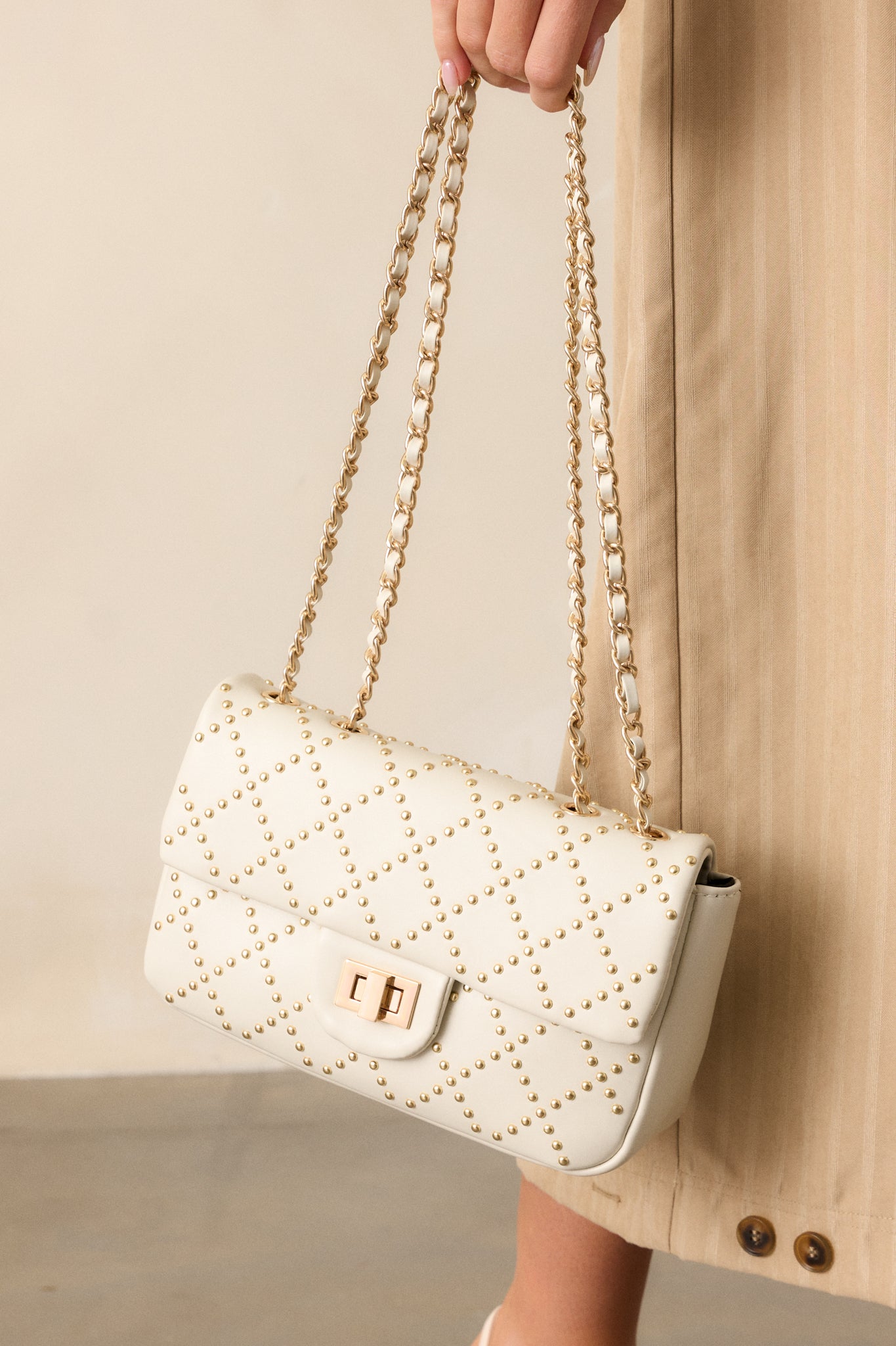 Full view of the ivory handbag, showcasing its quilted pattern with gold studs, gold twist-lock closure, and dual ivory and gold chain handles.