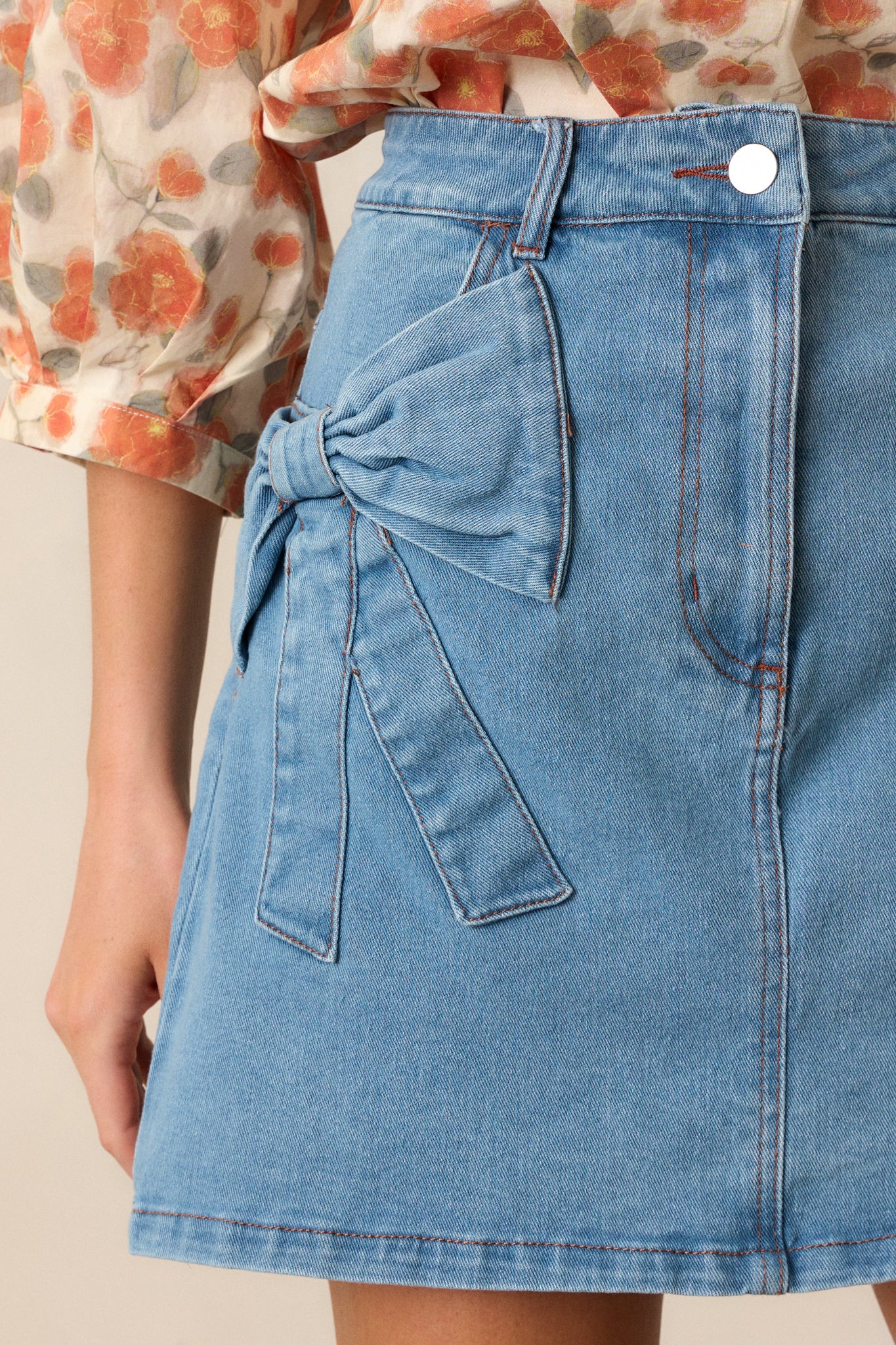 Close-up of the hip bow detailing and brown stitching on the light wash mini skirt, showing intricate fabric accents.