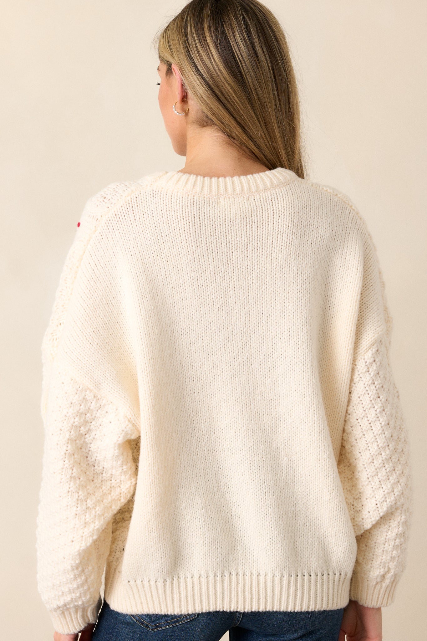 Back view of the oversized ivory sweater, showing a mix of cable knit patterns with subtle red and green thread accents and ribbed hem.