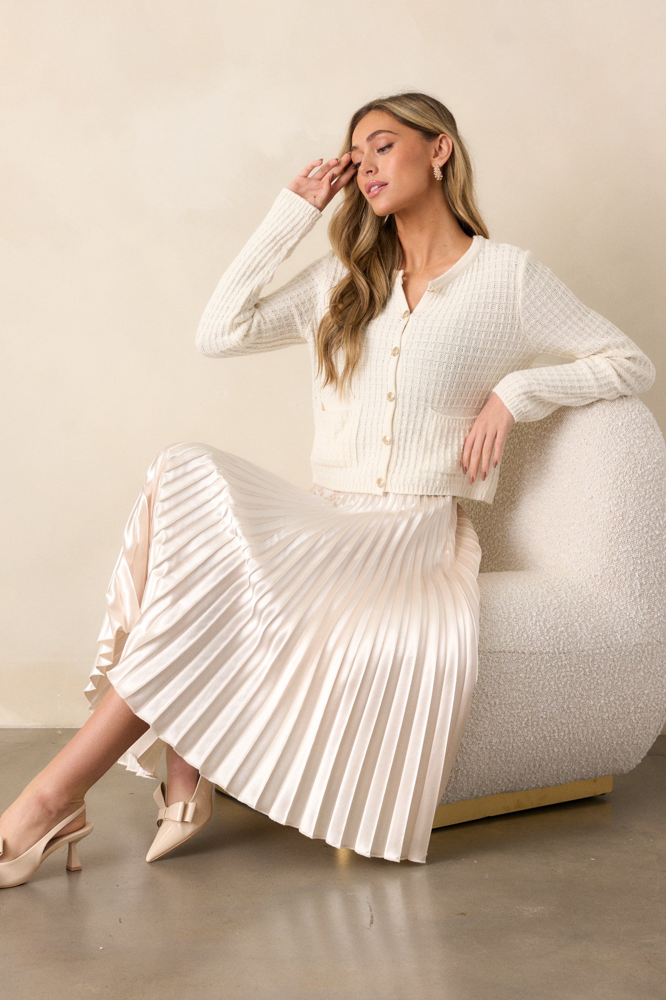 Seated, full length view of an ivory midi skirt featuring a high waisted design, an elastic insert at the back of the waist, a discrete side zipper, and heavy pleating throughout.