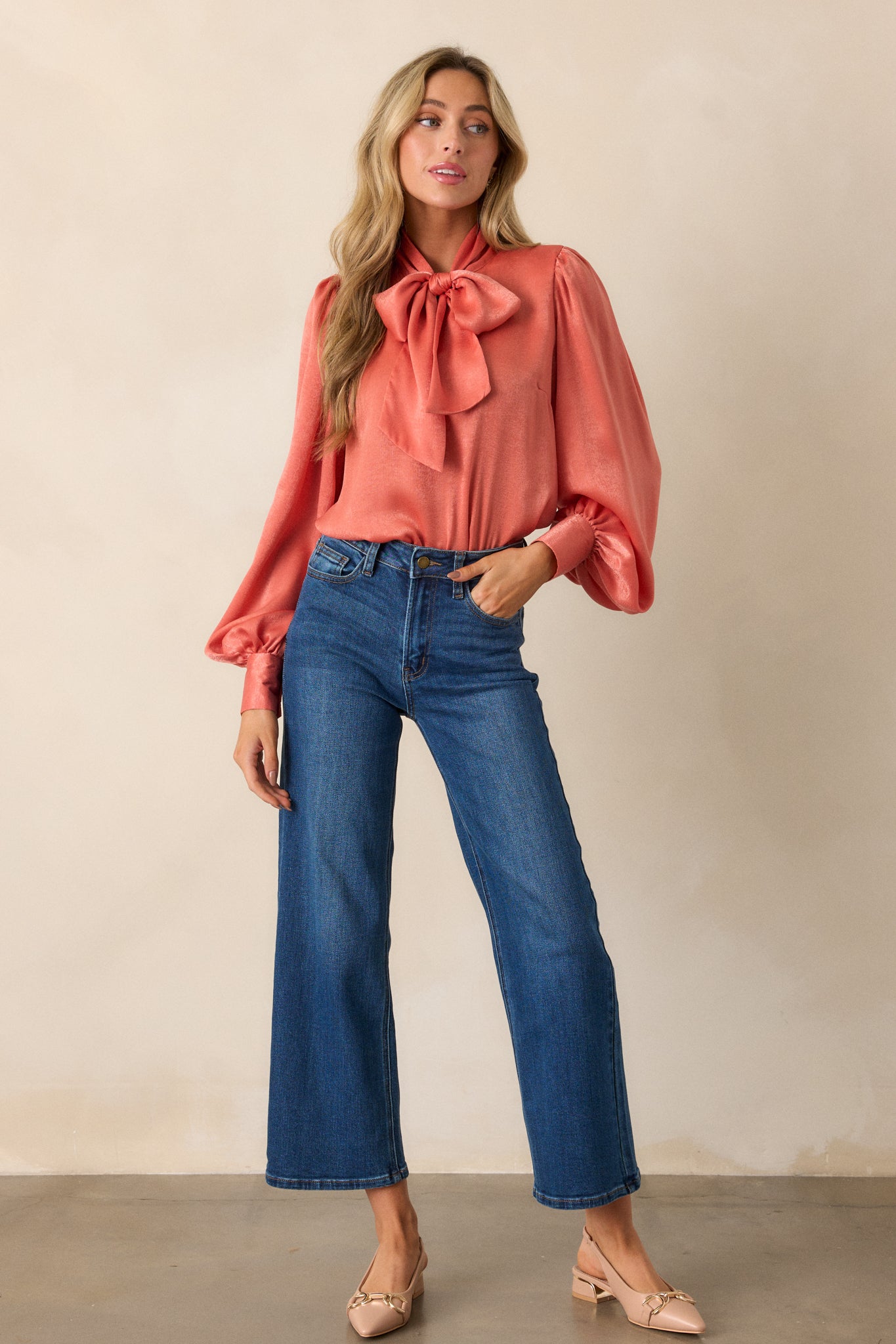 Full length view of a pink coral top with a high neckline, adjustable self tie, flowy balloon sleeves with two buttons at the cuff, and a relaxed fit.