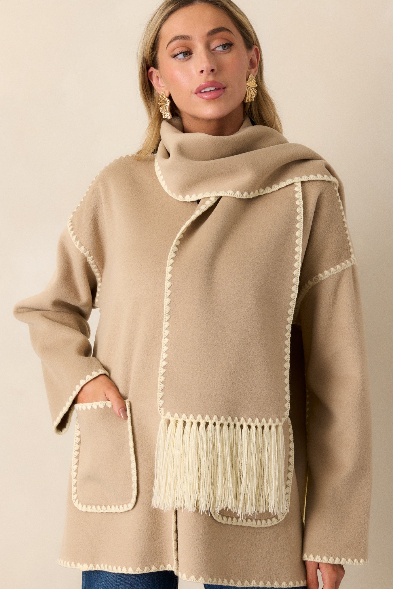 This Taupe Coat features a rounded neckline, functional button front, blanket stitch hems, functional front pockets, long sleeves with blanket stitch detailing and a blanket stitch scarf with fringe detailing.