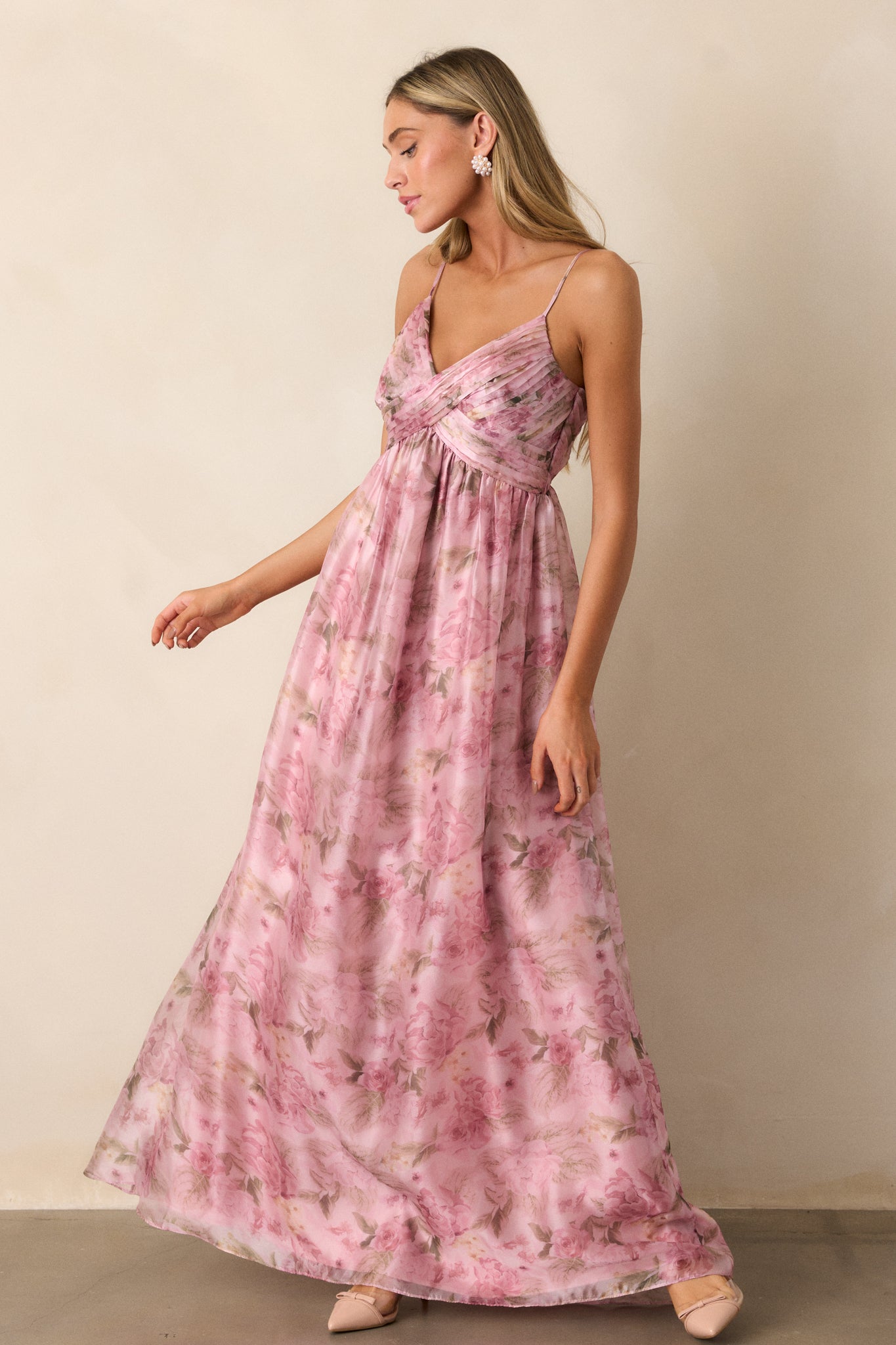 Front view of a pink dress featuring a v- neckline, empire waist, cross pleated detailing on the top, flowing skirt, an elegant floral design throughout, and spaghetti straps.