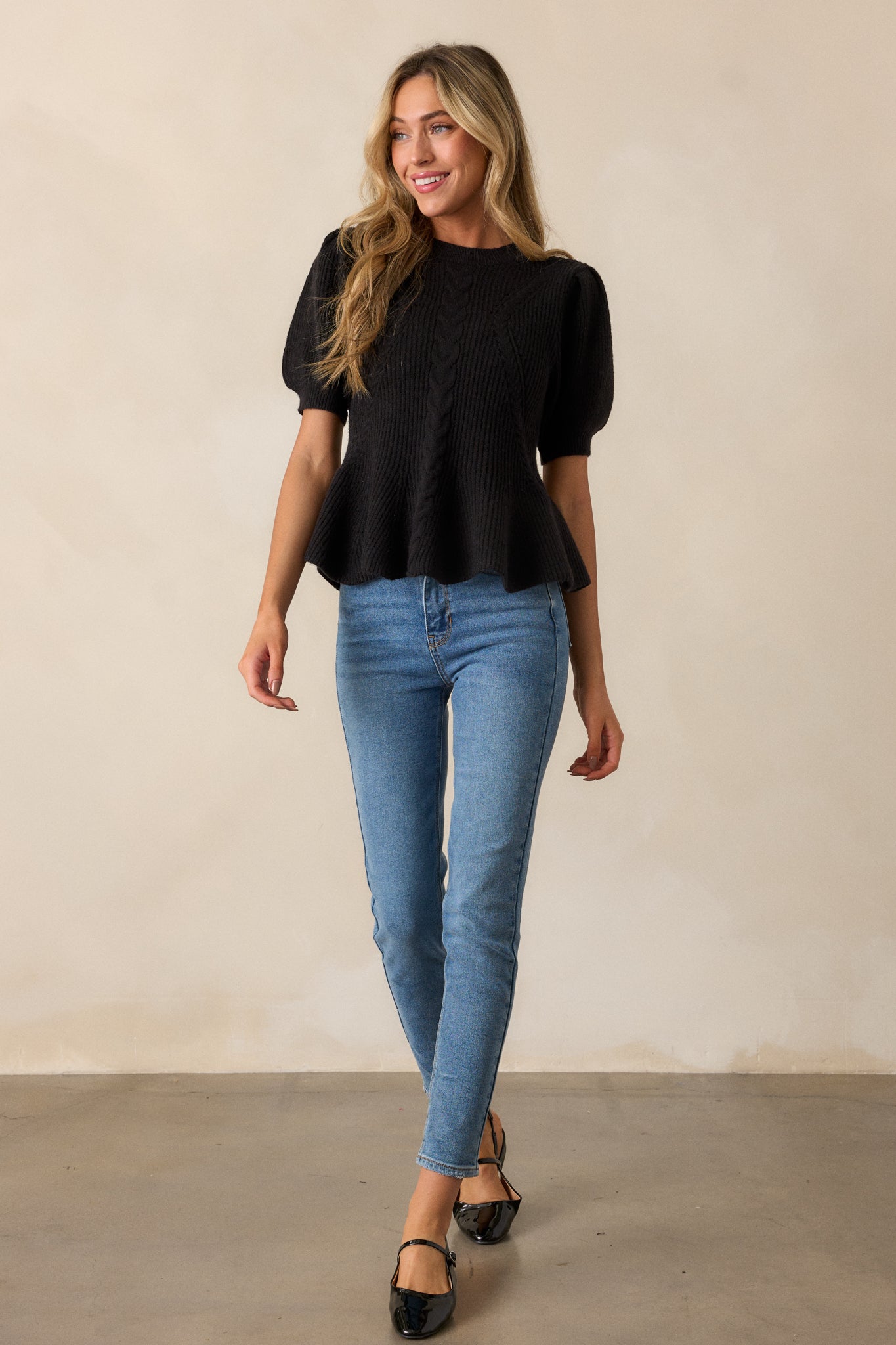 A black sweater with a rounded ribbed neckline, featuring braided knit detailing, a peplum fit, and puff sleeves with ribbed cuffs.