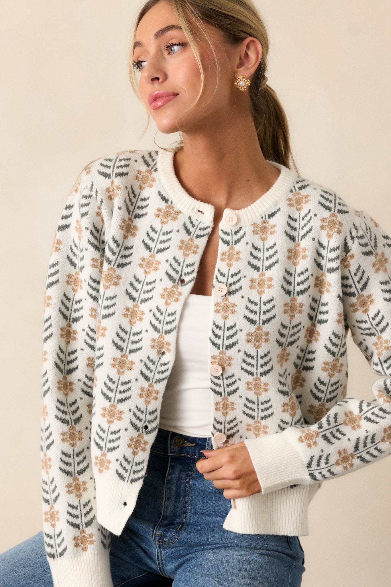 This ivory cardigan features a round neckline, a repeating pattern of delicate beige floral motifs with dark green leaf accents, ribbed hems and long sleeves.