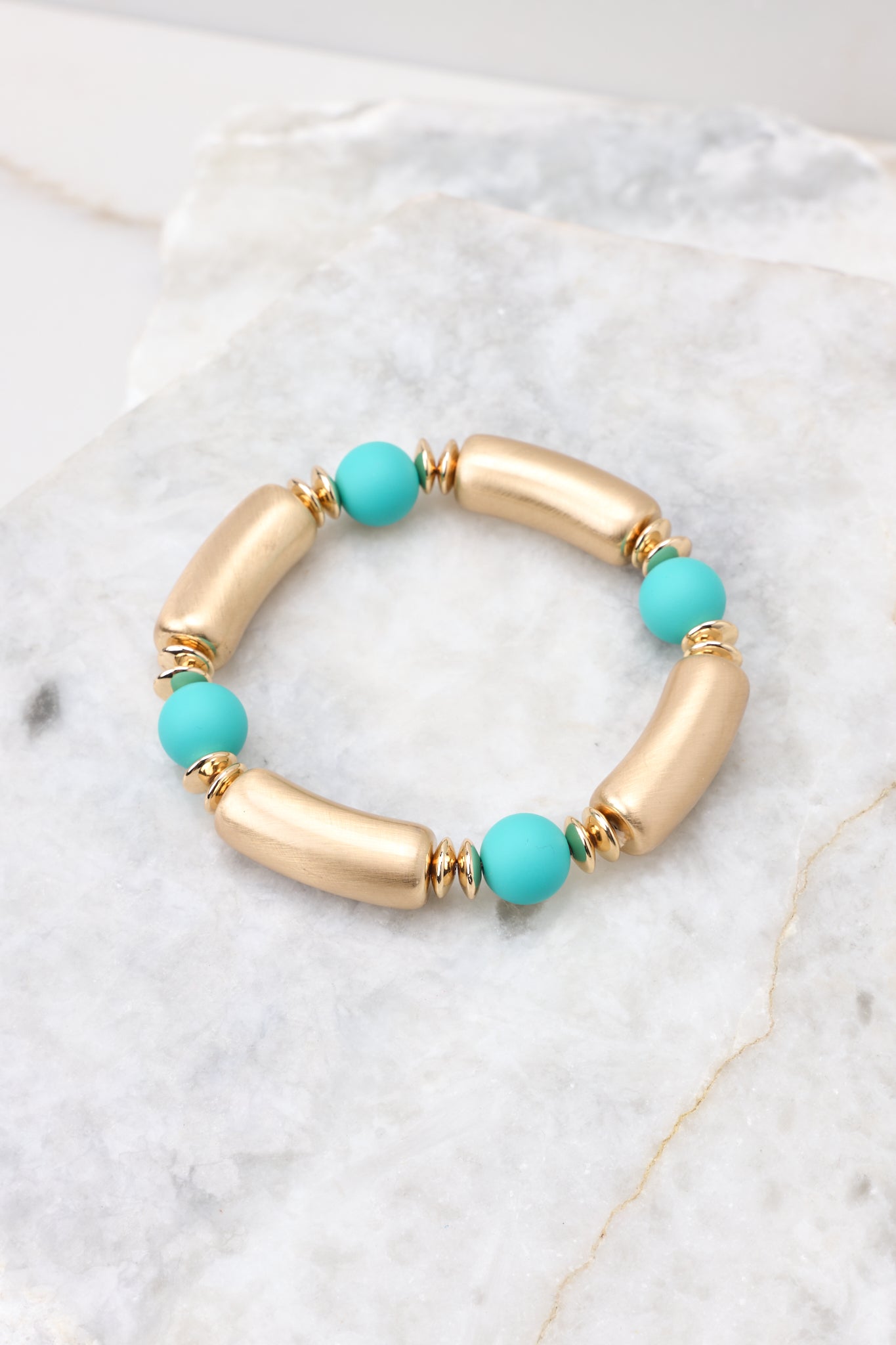 Detailed view of bracelet that features alternating gold and teal beads, with an elastic band underneath. 